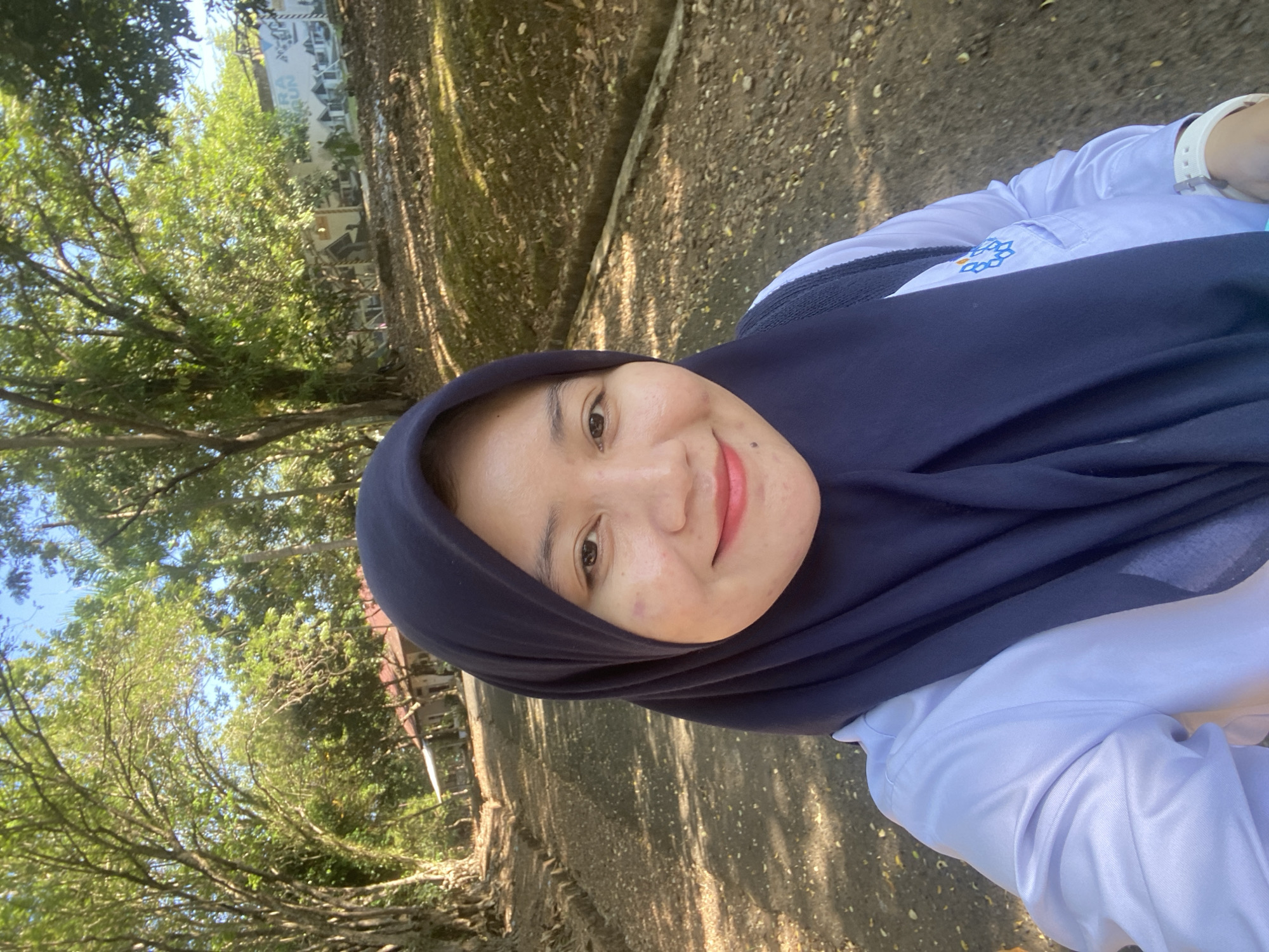 A young woman wearing a dark blue hijab and a light purple collared shirt, with a white smartwatch on her left wrist, is taking a selfie outdoors. She is smiling and has light makeup on. Behind her, there is a dirt path lined with trees and foliage, with a building and a billboard visible in the distance. The scene appears to be in a park or campus setting, possibly located in Banjarbaru, Indonesia. The sunlight suggests it is daytime.