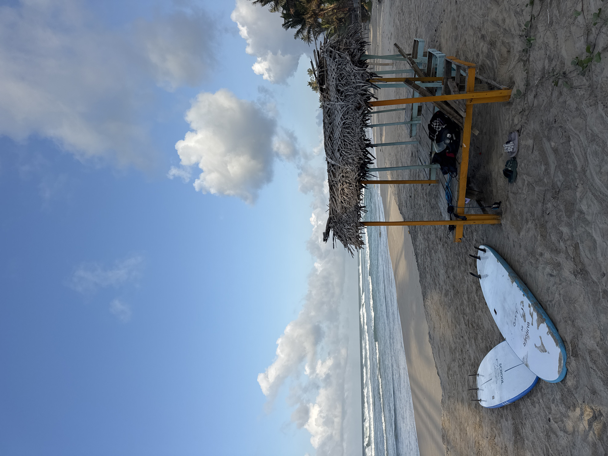 The image captures a serene beach scene in Kathaluwa, Sri Lanka, bathed in the soft light of what appears to be early morning or late afternoon. The foreground is dominated by a sandy beach, with two white surfboards resting side-by-side, their fins pointing upwards. The surfboards are a light blue and white, with visible markings on their surfaces. To the right, a rustic, open-sided shelter constructed with a thatched roof and yellow and blue wooden posts stands on the sand. Inside the shelter, there are wooden benches and several bags, including a dark-colored backpack and a helmet. A pair of dark green and light grey sandals are placed on the sand in front of the shelter. Beyond the beach, the ocean stretches out, with gentle waves rolling towards the shore. The water is a deep blue, contrasting with the lighter sandy beach. The sky above is a vibrant blue, dotted with scattered, fluffy white clouds. In the distance, palm trees line the edge of the beach, hinting at the tropical location. The overall atmosphere is peaceful and tranquil.