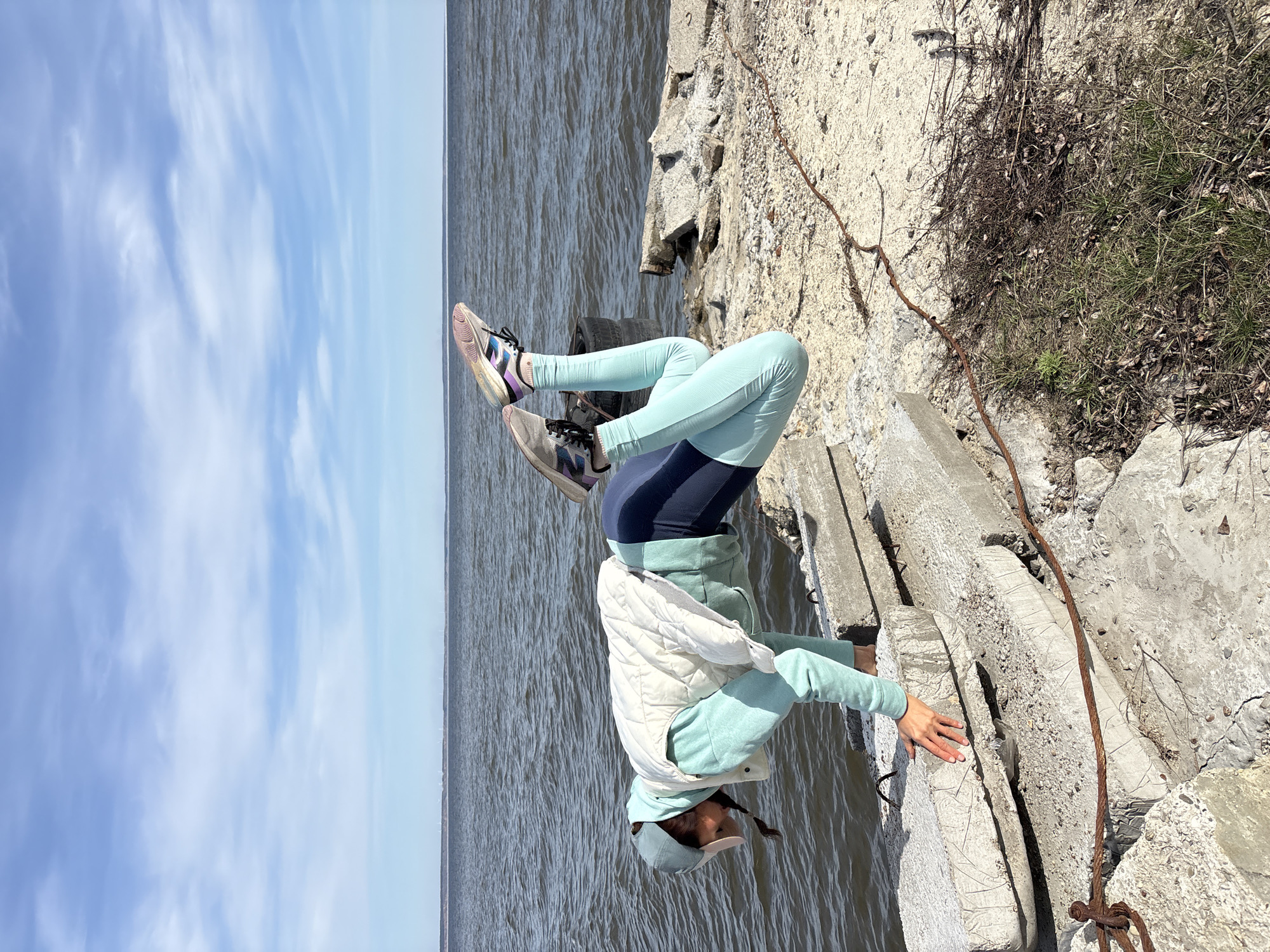 The image captures an individual performing an inverted physical pose, resembling a headstand or handstand, on concrete structures next to a large body of water. The person is positioned with their hands on rugged concrete blocks, their body inverted, and their legs bent upwards.
The subject is dressed in athletic attire, featuring light blue or mint green leggings and a matching long-sleeved top, overlaid with dark blue shorts and a light cream-colored quilted vest. They wear a light blue baseball cap and sneakers with a multi-colored design including grey, black, purple, and pink accents.
The setting is an outdoor, waterfront location, likely an embankment or riverbank. The foreground is dominated by rough, cracked concrete slabs and blocks, with some dry grass, weeds, and debris growing in between. A rusty, thick metal cable or rebar is visible, running along and embedded in the concrete. Beyond the immediate foreground, a wide expanse of water extends to the horizon, meeting a clear, bright blue sky with scattered white clouds. The scene is bathed in natural daylight, indicating midday or early afternoon.
Given the location context of Ulyanovsk, Russia, the body of water is presumably the Volga River. There is no visible text in the image.