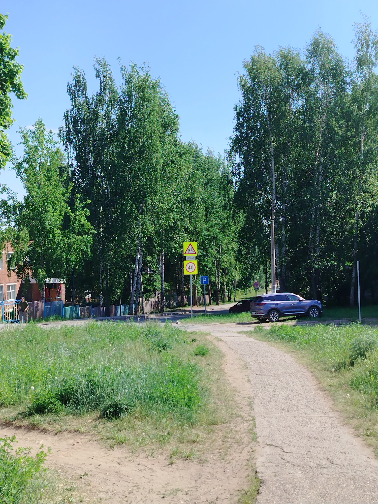 The image shows a street scene in Noviy, Russia. A worn, cracked asphalt path leads from the foreground towards a street intersection. The path is flanked by overgrown grass. At the intersection, a blue-gray SUV is parked on the side of the road, partially obscured by trees. A dark-colored car is parked behind the SUV. A signpost displays a pedestrian crossing warning symbol above a speed limit sign of 40, and a parking symbol. The road is lined with birch trees. In the background, a building is visible, partially obscured by trees and a colorful fence. The sky is clear blue. The scene is lit by bright daylight.