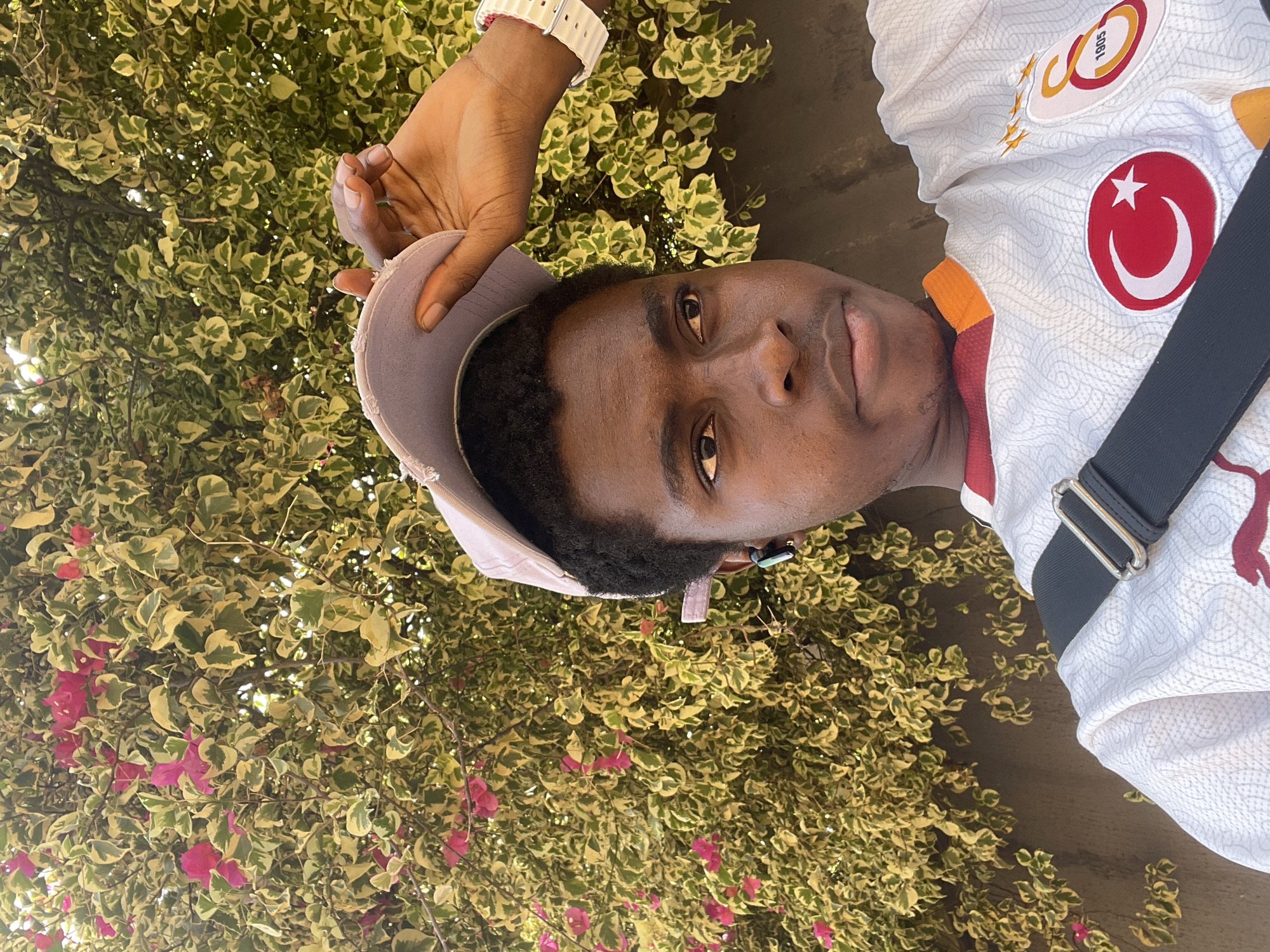 A young man stands outdoors, likely in Onitsha, Nigeria, judging by the local flora. He has dark skin and short, dark hair. The man is wearing a white jersey with a Turkish flag emblem. He is also wearing a light-colored baseball cap and a white wristwatch. He holds the brim of his cap with his right hand. A black strap is across his body, likely belonging to a bag. The background contains dense vegetation with green and yellow leaves, and some bright pink flowers.