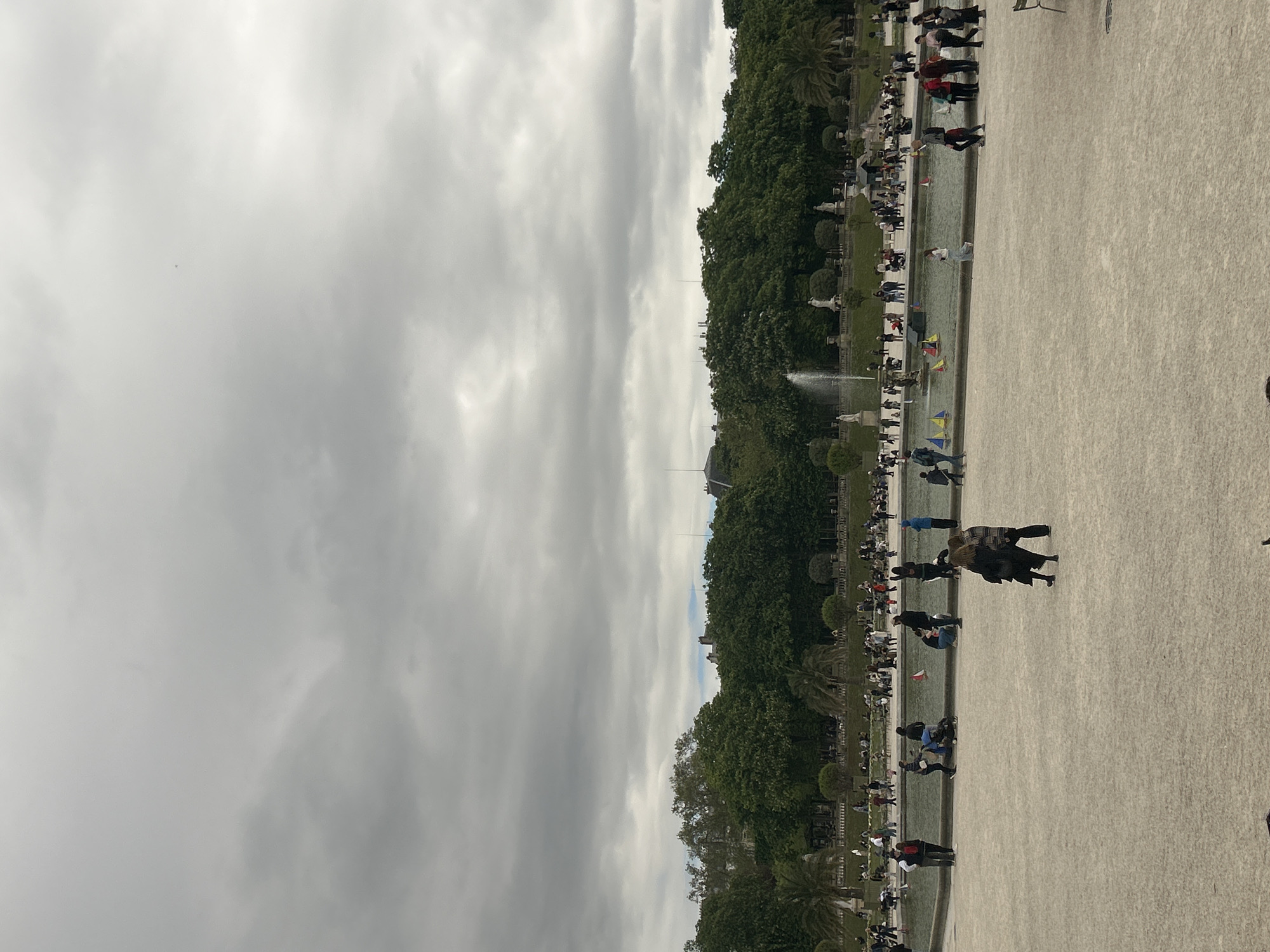 The image depicts the Jardin des Tuileries in Paris, France, on a cloudy day. The garden features a long, rectangular pool with a fountain in the distance, lined with trees, statues, and manicured hedges. The pool is filled with miniature sailboats. Many people are visible along the edges of the pool, some walking and others sitting. The ground is a light-colored gravel. The sky is overcast with gray clouds.