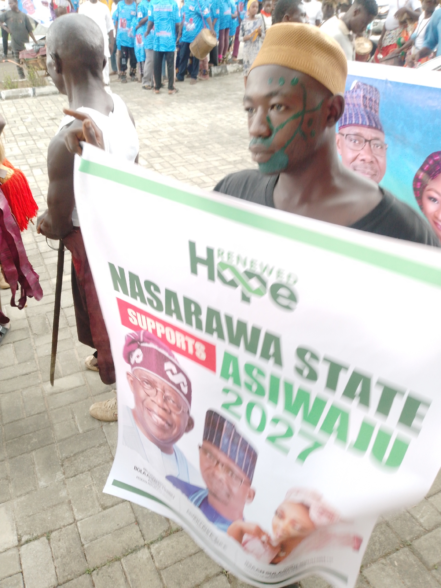 The image captures a political gathering in an unidentified location in Unknown, Unknown. A man with green markings on his face and a yellow cap holds a banner that reads "Renewed Hope NASARAWA STATE Supports ASIWAJU 2027." The banner also features images of political figures. Surrounding the man are other people, including a person holding a walking stick and several individuals wearing blue shirts and playing drums. The scene takes place on a paved surface, suggesting an urban environment.