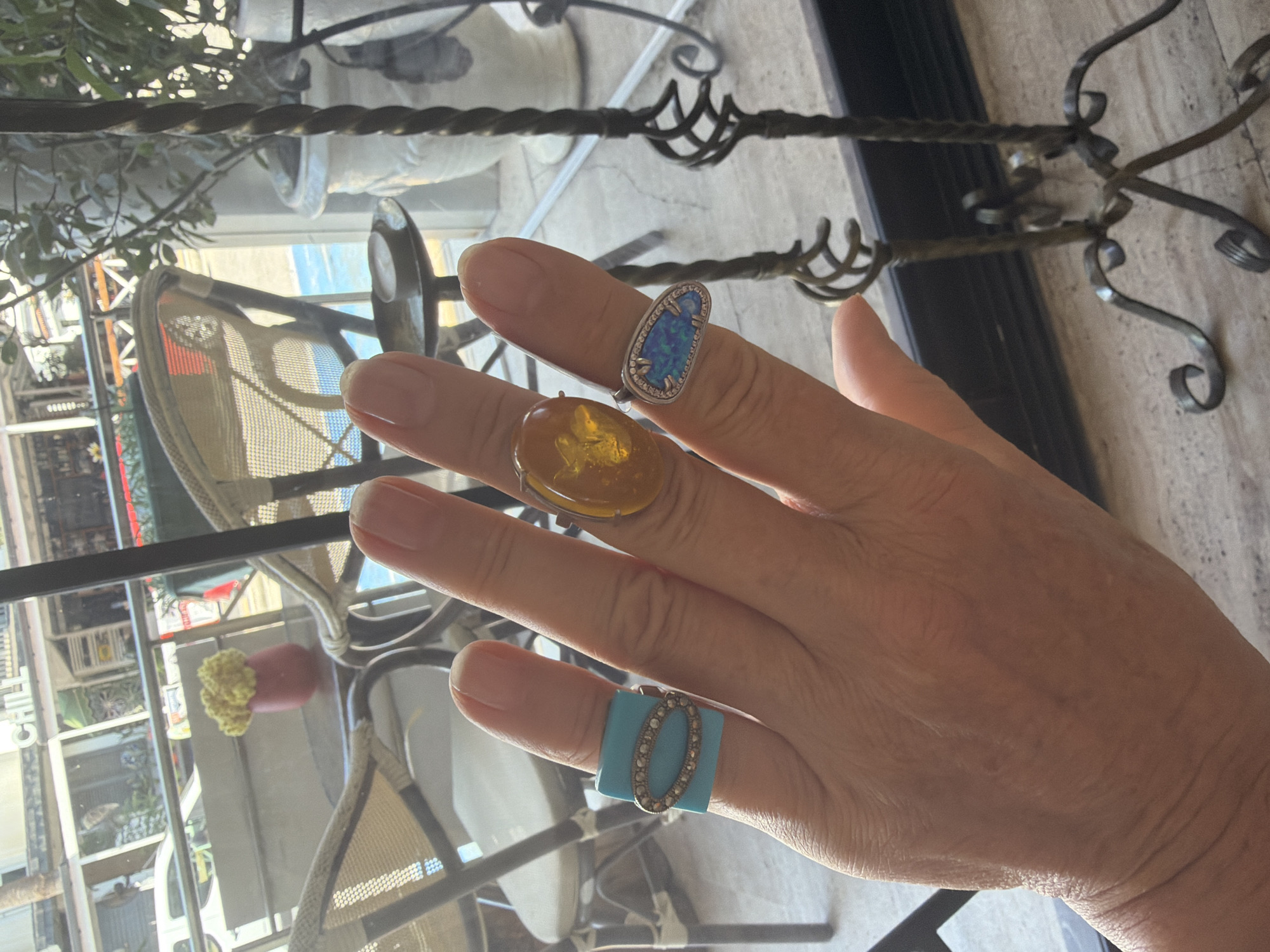A hand with three rings is shown in the foreground. From left to right, the rings are a turquoise rectangular stone with small diamonds in an oval shape, a large oval amber stone, and a silver ring with an oval blue and white opal stone surrounded by small diamonds. In the background, there are outdoor cafe tables and chairs with people blurred in the distance. The scene appears to be in an outdoor seating area of a cafe or restaurant in Marmaris, Turkey, judging by the style of the furniture and the general ambiance.