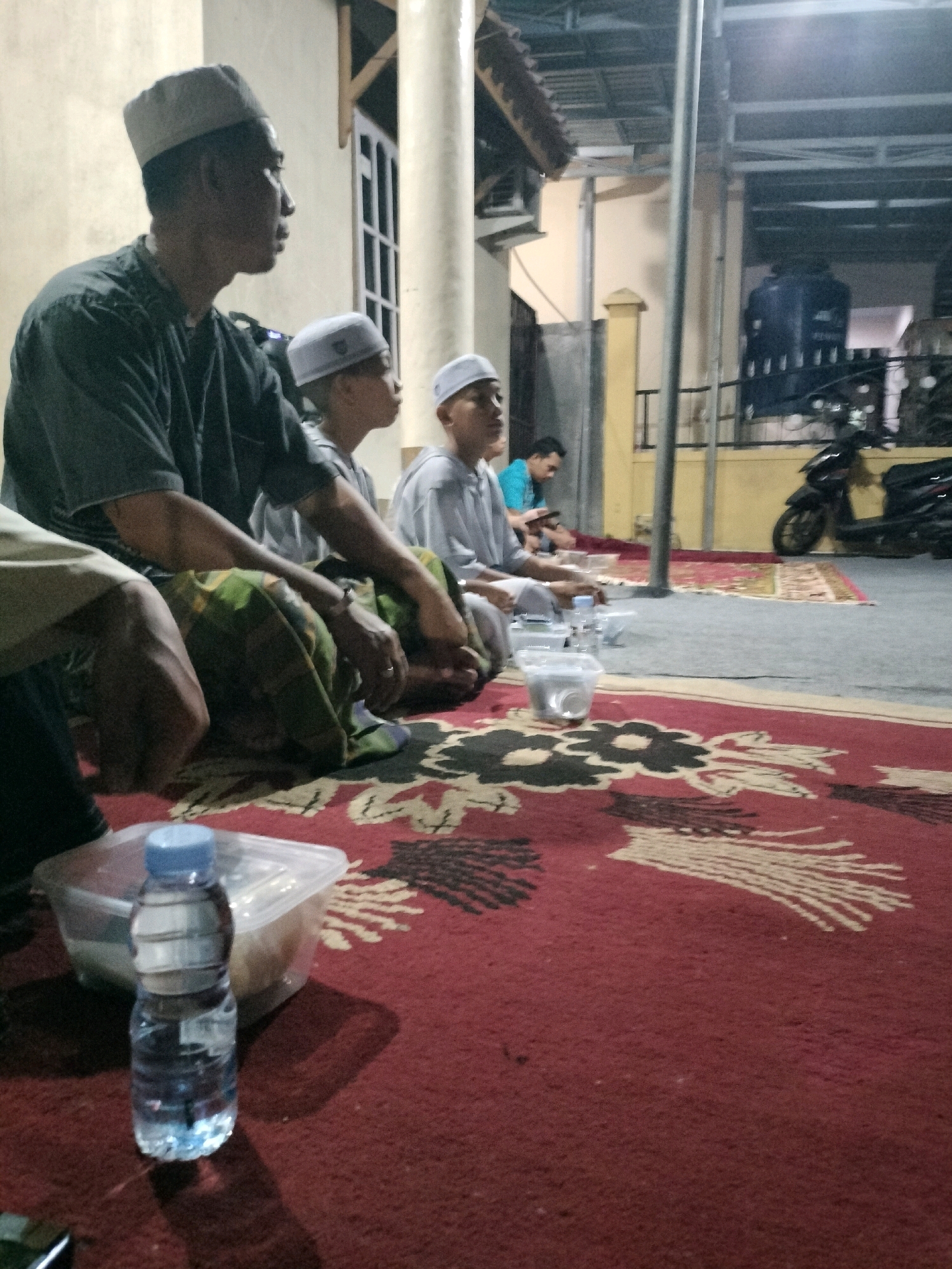 In Pembataan, Indonesia, an outdoor gathering takes place in the evening. Three men, wearing traditional Islamic attire including white caps, are seated on a red carpet with a floral pattern. An older man in a dark shirt and a younger man in a light blue shirt are in the foreground, while another young man in a light gray shirt sits behind them. A plastic bottle of water and a clear plastic container are placed on the carpet in front of the older man. In the background, a motorcycle is parked near a building with a large blue water tank. The scene appears to be a casual gathering, possibly a family or community event.