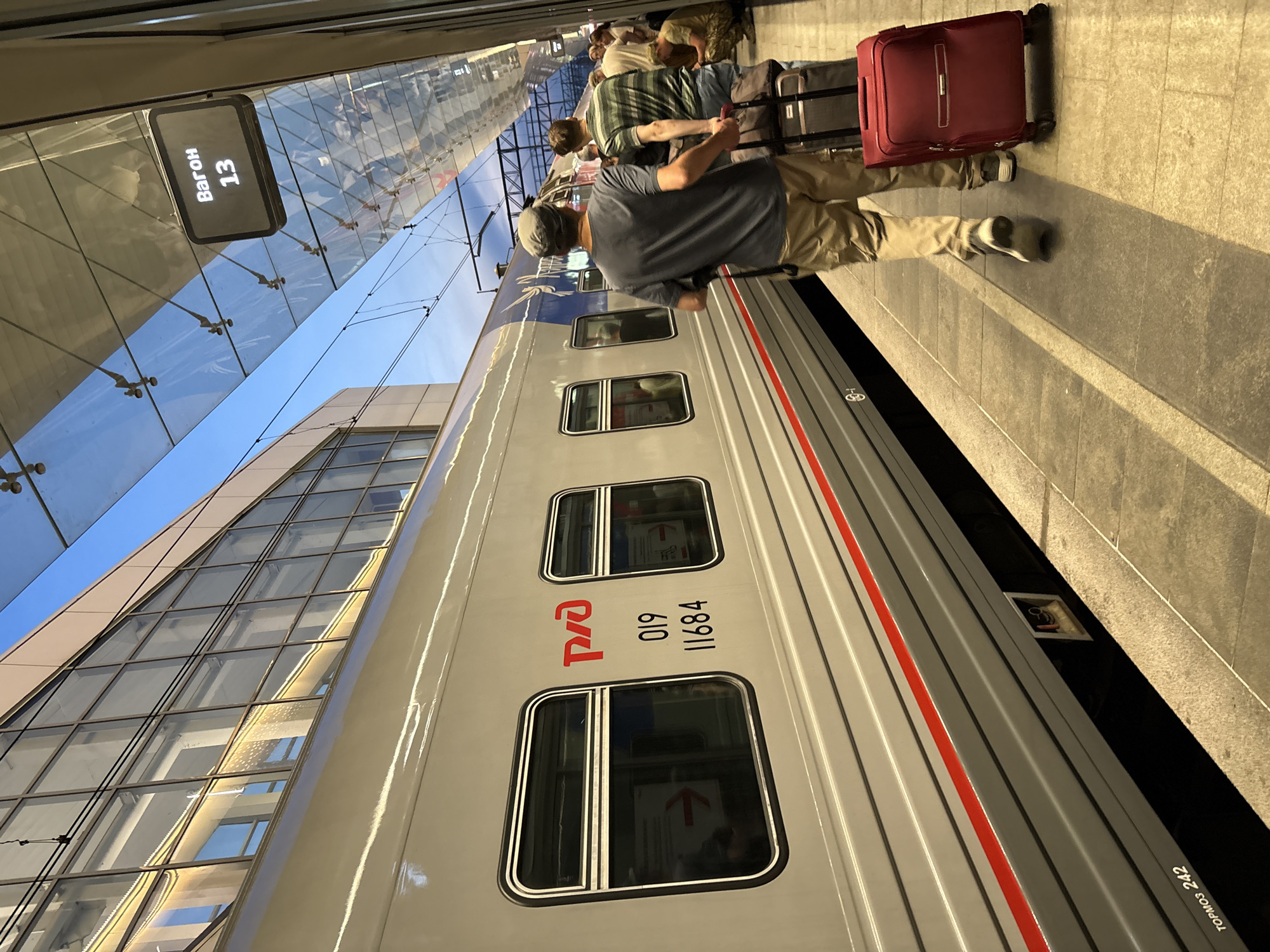 The image depicts a train at a station platform. A modern train car, predominantly gray with a thin red stripe running horizontally along its side, is the main subject. Two men are walking away from the camera on the platform. The man in the foreground is wearing a gray t-shirt, khaki pants, and a gray cap, and he is pulling a black rolling suitcase. The man behind him wears a striped green and blue shirt and is also pulling a black rolling suitcase. Red luggage is also visible on the platform. Above the platform, a glass canopy with exposed metal supports covers the area, and an illuminated digital sign displays "Вагон 13" (Car 13) in white text on a black background. The building to the left features a large, modern glass facade. In the background, other passengers and a few parked vehicles are partially visible. The lighting suggests it is either early morning or late afternoon. The presence of the "РЖД" logo (Russian Railways) on the train confirms its origin in Russia. The specific location within Moscow can be inferred as a major railway station such as Leningradsky, Yaroslavsky, or Kazansky, given the scale and modern architecture.