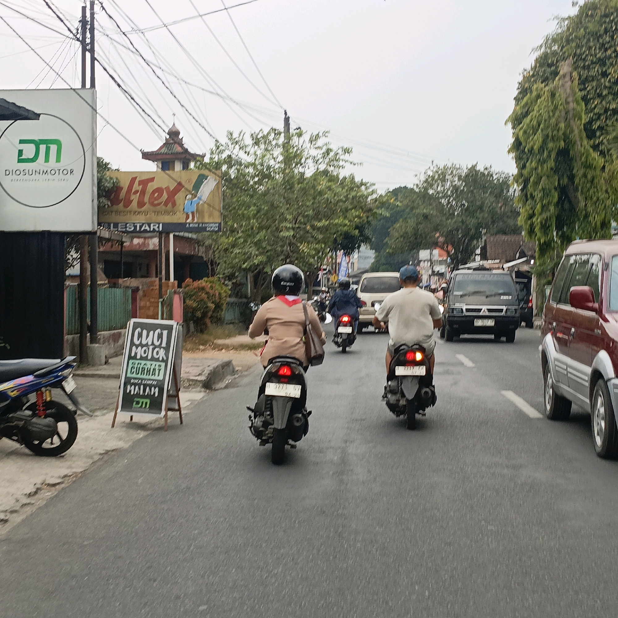 The image depicts a street scene in Salatiga, Indonesia. In the foreground, two people are riding motorcycles. The motorcycle on the left has a blue and yellow design. The rider of the motorcycle in the center is wearing a light brown uniform with a red scarf and a black helmet, and is carrying a brown bag over their shoulder. The rider of the motorcycle to the right is wearing a light-colored shirt and a blue cap. The street is paved and has white lane markings. On the left side of the road, there is a sign for "DIOSUNMOTOR". Further down the street, there is a large billboard with the text "Al-Tex" and an illustration of a person holding a paint can and roller. There are also other vehicles visible, including a dark-colored van and a red SUV. Trees and buildings line the street, and there are numerous power lines overhead. In the lower left corner, a sign for "CUCI MOTOR" (motorcycle wash) is visible, indicating services offered.