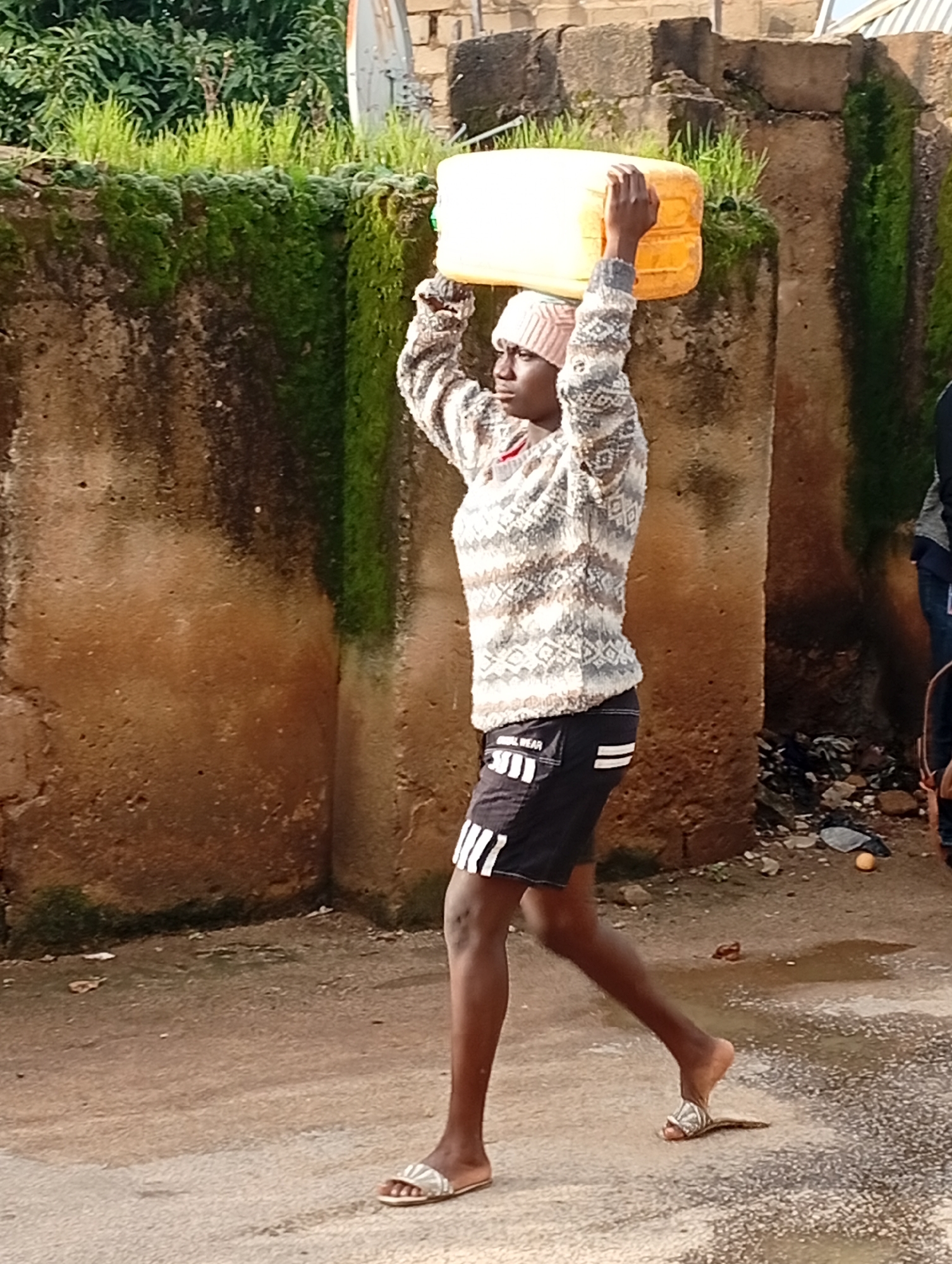 A young person, possibly a teenager, walks across a paved path in Jos, Nigeria, carrying a large yellow plastic water container on their head. They are wearing a patterned sweater, dark shorts with white stripes, a pink beanie, and silver sandals. The background consists of a textured, brownish wall covered in patches of moss and greenery, with some brickwork visible above. The ground is dusty and uneven, with debris scattered in places. The lighting suggests it is daytime.