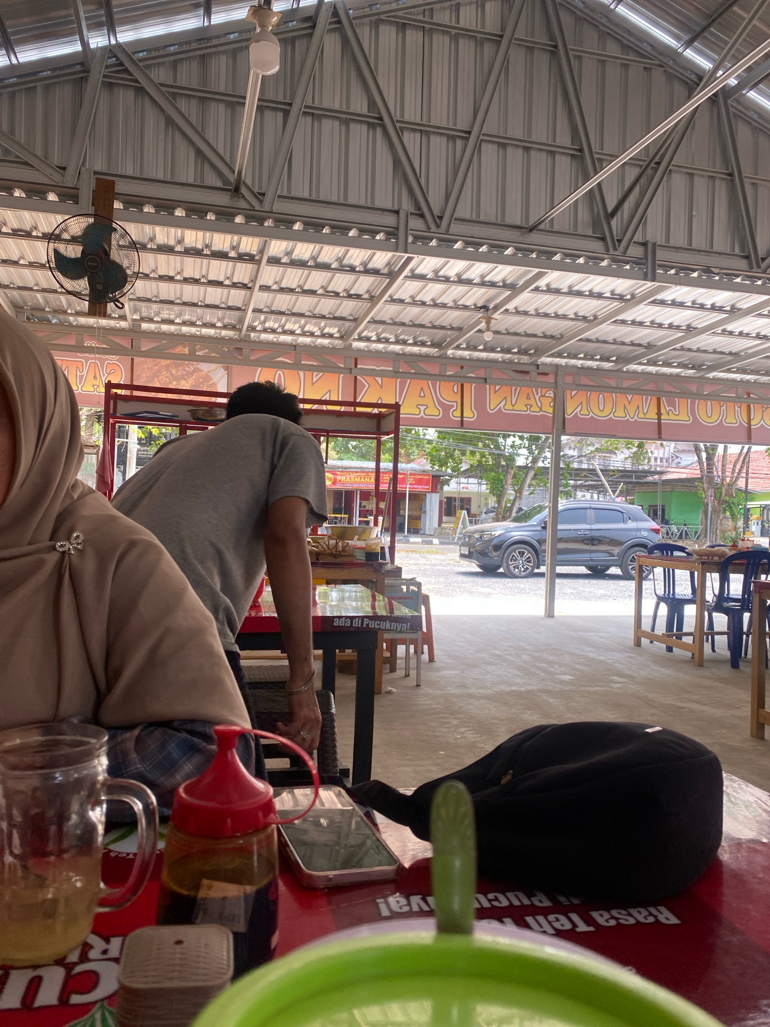 The image depicts an outdoor dining area of a restaurant in Banjarbaru, Indonesia. A person wearing a light brown hijab is seated on the left, with their left side facing the camera. To their right, a person wearing a grey t-shirt is standing and facing away from the camera, appearing to be interacting with something behind a red-colored table. In the foreground, a red table is visible, laden with various items including a glass mug, a red-capped condiment bottle, a smartphone, and a black bag. In the background, a dark grey SUV is parked on a paved area. The establishment has a metal roof supported by a metal frame, and signage with Indonesian text is visible on the building in the background. The overall environment suggests a casual, open-air eatery.