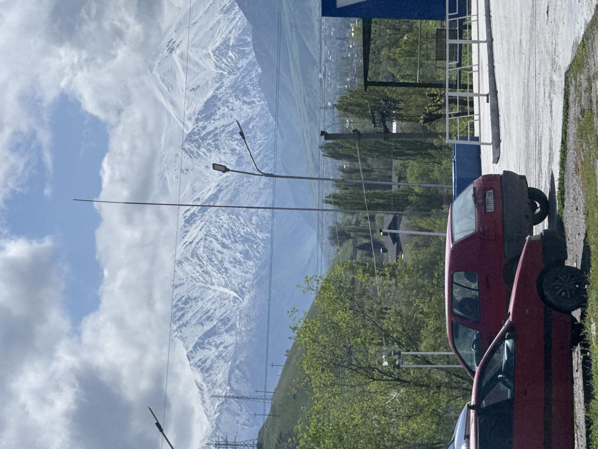 This daytime image captures a vast landscape featuring snow-capped mountains under a partly cloudy sky, juxtaposed with elements of human infrastructure and activity in what appears to be the foothills of Bishkek City, Kyrgyzstan.
In the background, majestic snow-covered mountains dominate the horizon, showcasing rugged terrain and remnants of winter snowmelt. Below the snow line, gentler green slopes suggest the arrival of spring or early summer. The sky above is a mix of clear blue patches and expansive white and grey clouds.
In the midground and foreground, signs of human settlement are evident. Numerous power lines and utility poles crisscross the scene, with several modern streetlights extending upwards. Lush green trees, indicative of fresh spring foliage, are abundant, particularly on the right side of the frame. A paved road or parking area occupies the lower right, with patches of grass and dirt along its edge. Partial views of buildings, featuring dark blue sections and white railings, are visible towards the right.
Two red cars are parked on the paved surface in the foreground. The vehicle closer to the viewer is partially obscured, while the car behind it is clearly visible, appearing to be an older model hatchback. A visible license plate on the rear of this car displays "01 724CAA" with "KG" underneath, confirming its origin from Kyrgyzstan.
The scene is brightly lit by daylight, with soft shadows. There is no specific activity depicted; rather, it’s a static view emphasizing the dramatic natural scenery meeting a developed area. The overall impression is one of natural grandeur meeting urban edge in a temperate climate.