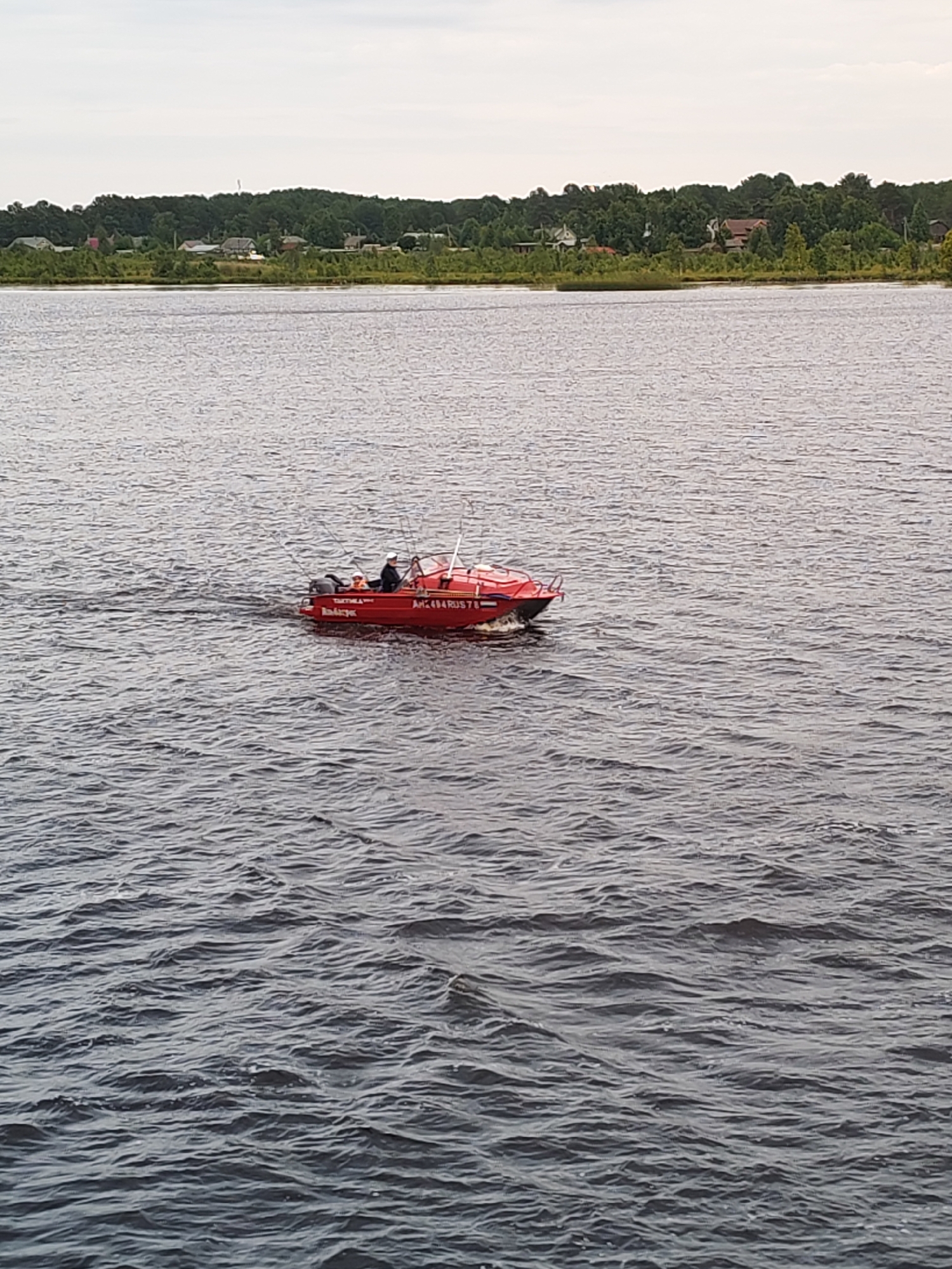 A red boat with three people on board is moving across a body of water, likely a river, near the city of Shal'skii, Russia. The boat has text written on its side, including "AH2 494 RUS 78". The water is choppy, and in the background, a tree-lined shore with scattered houses can be seen under a cloudy sky.