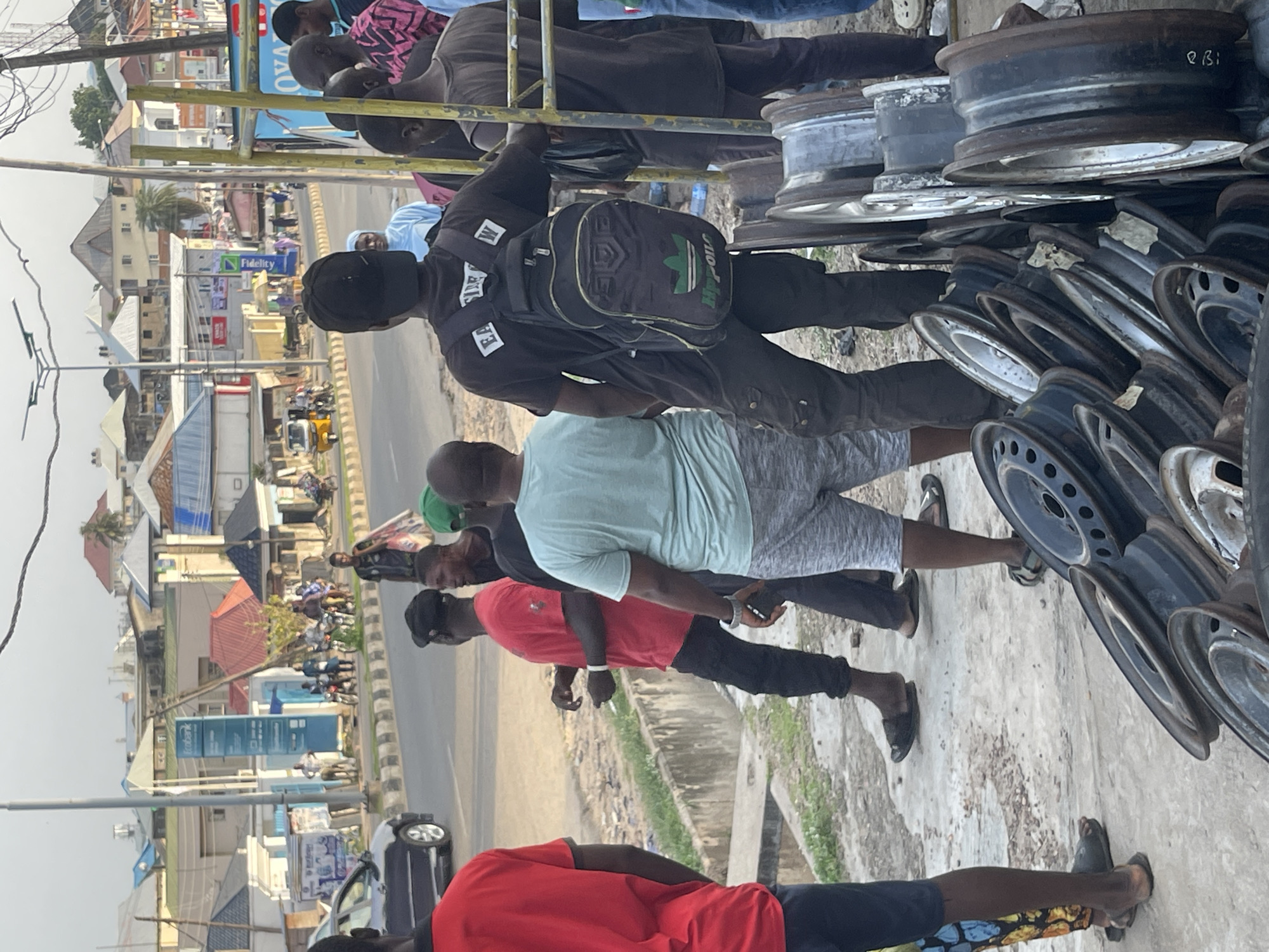 The image shows a group of people gathered near a roadside stall where car rims are displayed. Several men are standing and looking at the rims, which are stacked and arranged for sale. Some of the men appear to be potential buyers or vendors.
The scene is set outdoors, likely on a street in a commercial area, given the presence of buildings and what appears to be a road. The time of day seems to be daytime, with natural light illuminating the scene. The weather appears to be clear, with no signs of rain or strong wind.
One individual in the foreground is wearing a red shirt and dark pants. Another person is in a light blue t-shirt and grey shorts. A person further back is wearing a black t-shirt and dark pants, carrying a backpack with an Adidas logo. There are also other individuals visible in the background, some of whom are wearing more traditional clothing.
The car rims are the main objects in focus, with various sizes and types visible. They are made of metal and appear to be used or for sale at a relatively low cost. Some rims are rusty, suggesting they might be second-hand.
There is some visible text in the background, including a blue sign with the word "Fidelity" and another sign with the word "Ecobank". These indicate the presence of financial institutions nearby, suggesting an urban or semi-urban commercial setting. The overall atmosphere seems relaxed, with people casually interacting and browsing the merchandise.