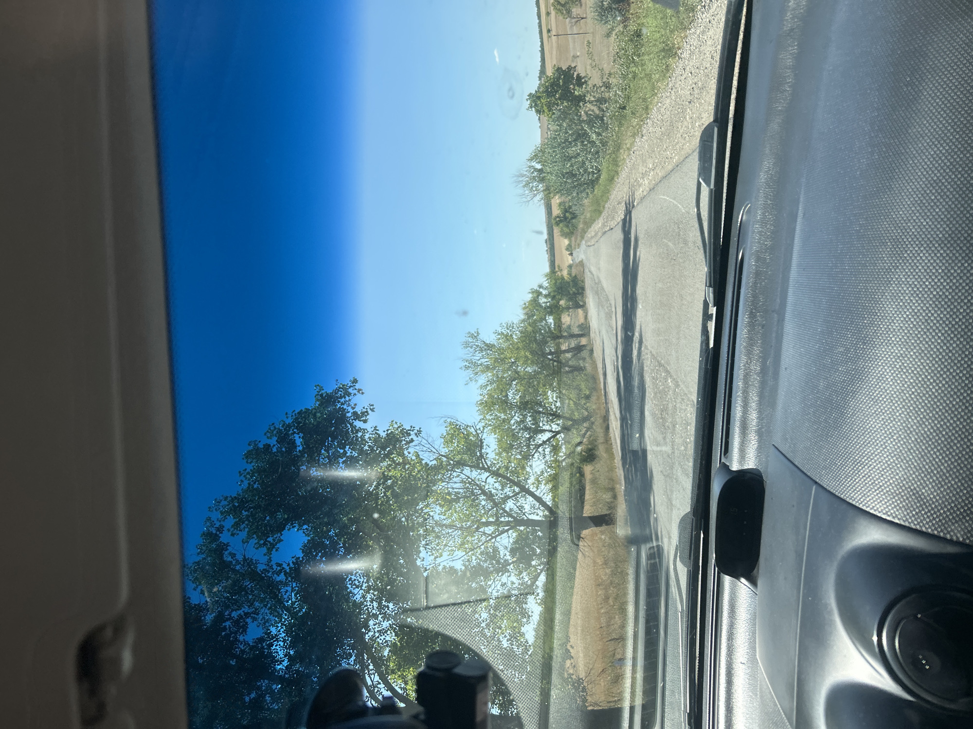 The image is a first-person perspective from inside a vehicle, looking through the windshield at a rural road. The sky is a clear, bright blue. The road is paved and appears to be winding into the distance. On either side of the road are fields of dry grass or crops, with scattered trees and bushes. There are some light reflections on the windshield. The dashboard of the car is visible in the foreground, with a small digital display showing "75". The scene appears to be a sunny day in a rural area, possibly the outskirts of Cioc-Maidan, Moldova, given the dry landscape and vegetation.