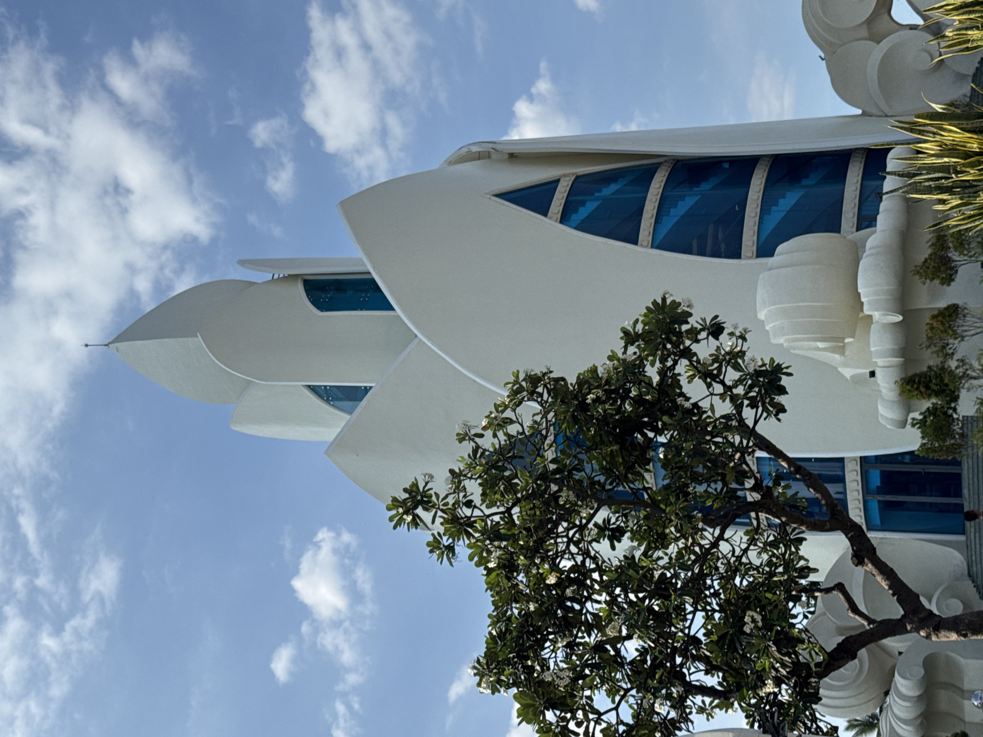 A white, futuristic building with a flower-like design stands against a bright blue sky with scattered white clouds. The building features sharp, white curves and large, triangular, blue-tinted windows that reveal spiral staircases inside. In the foreground, a leafy green tree with small white flowers partially obscures the building's lower section. To the right and left of the building, there are abstract, wave-like white sculptures. The scene appears to be an exterior shot of the One Pillar Pagoda (Chua Mot Cot) in Nha Trang, Vietnam, a replica of the famous Hanoi structure.