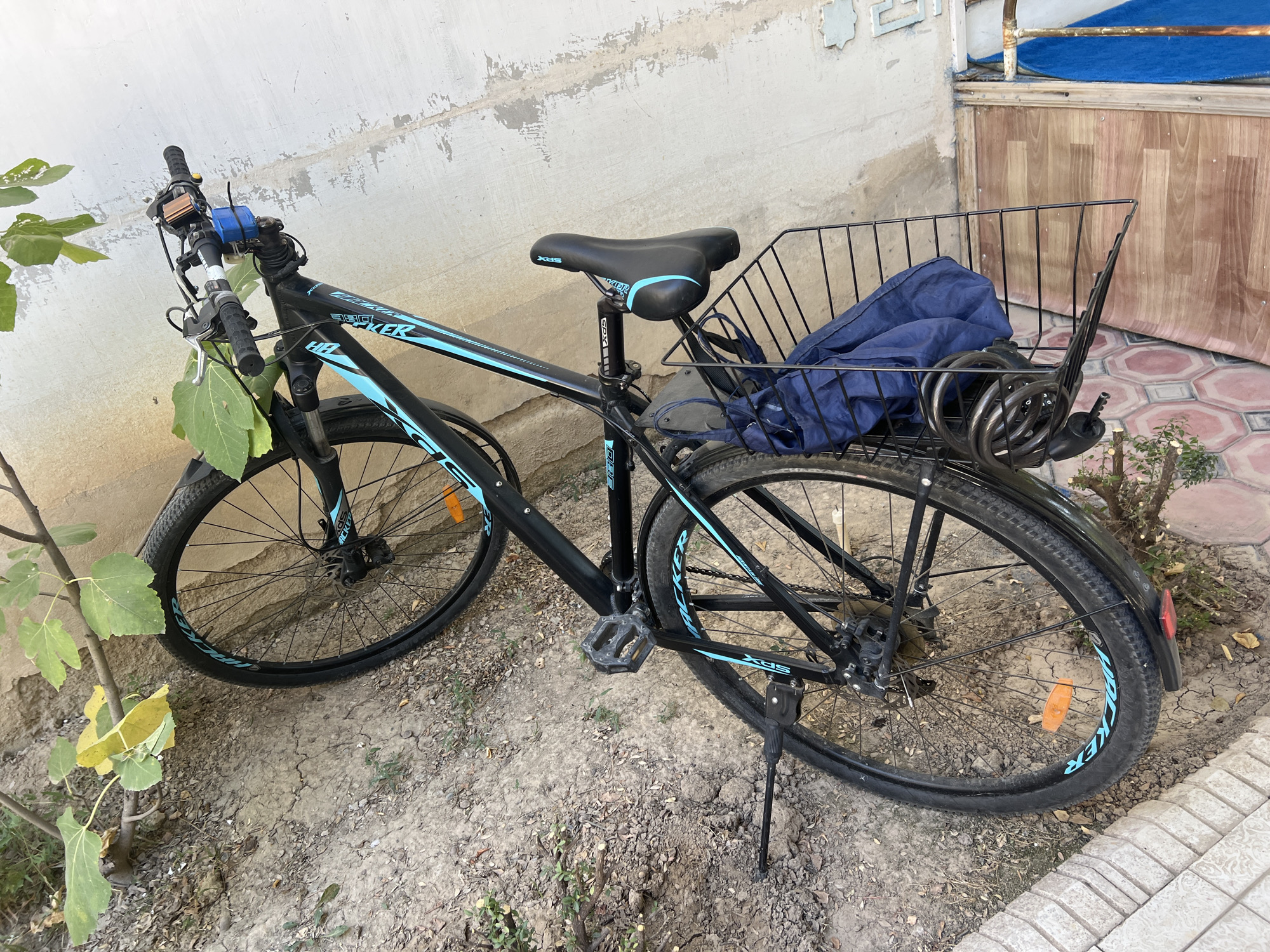 A black bicycle with blue accents is parked outdoors, leaning on its kickstand. The bicycle has a front basket containing a folded blue tarp and a coiled black bike lock. The seat is black with blue trim. The handlebars are equipped with a bell, brake levers, and what appears to be a small battery-powered light. The bicycle is positioned on dry, uneven ground with some scattered leaves and small plants. In the background, a textured, light-colored wall and a wooden structure with patterned tiling are visible. The setting is outdoors, possibly a courtyard or alleyway, in an urban environment.