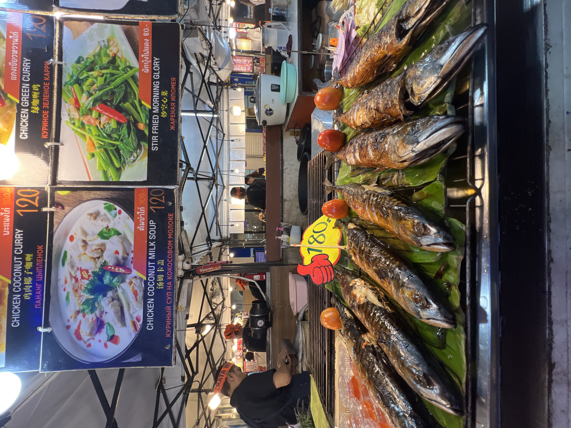 This is a photograph taken at a food stall in Karon, Thailand, likely in the evening or at night due to the artificial lighting. The scene depicts a vibrant street food market, with various dishes displayed for sale.
In the foreground, there are several grilled fish laid out on banana leaves. To the left, menus are displayed, showcasing dishes like "CHICKEN COCONUT CURRY" and "CHICKEN GREEN CURRY" with prices indicated in numbers and local currency. Another menu features "CHICKEN COCONUT MILK SOUP" and "STIR FRIED MORNING GLORY." A prominent yellow sign with a red hand symbol displays the price "180."
People are visible in the background, with one individual wearing a black shirt and a headband that reads "KATA," suggesting they might be working at the stall or a nearby establishment. Other individuals can be seen tending to the food or browsing the market. The setting is outdoors, with the stall under a temporary structure, common in street food markets. The overall impression is a bustling and lively atmosphere, characteristic of Thai night markets. The food items are attractively presented, with fresh ingredients and appealing colors, indicating a focus on local cuisine.