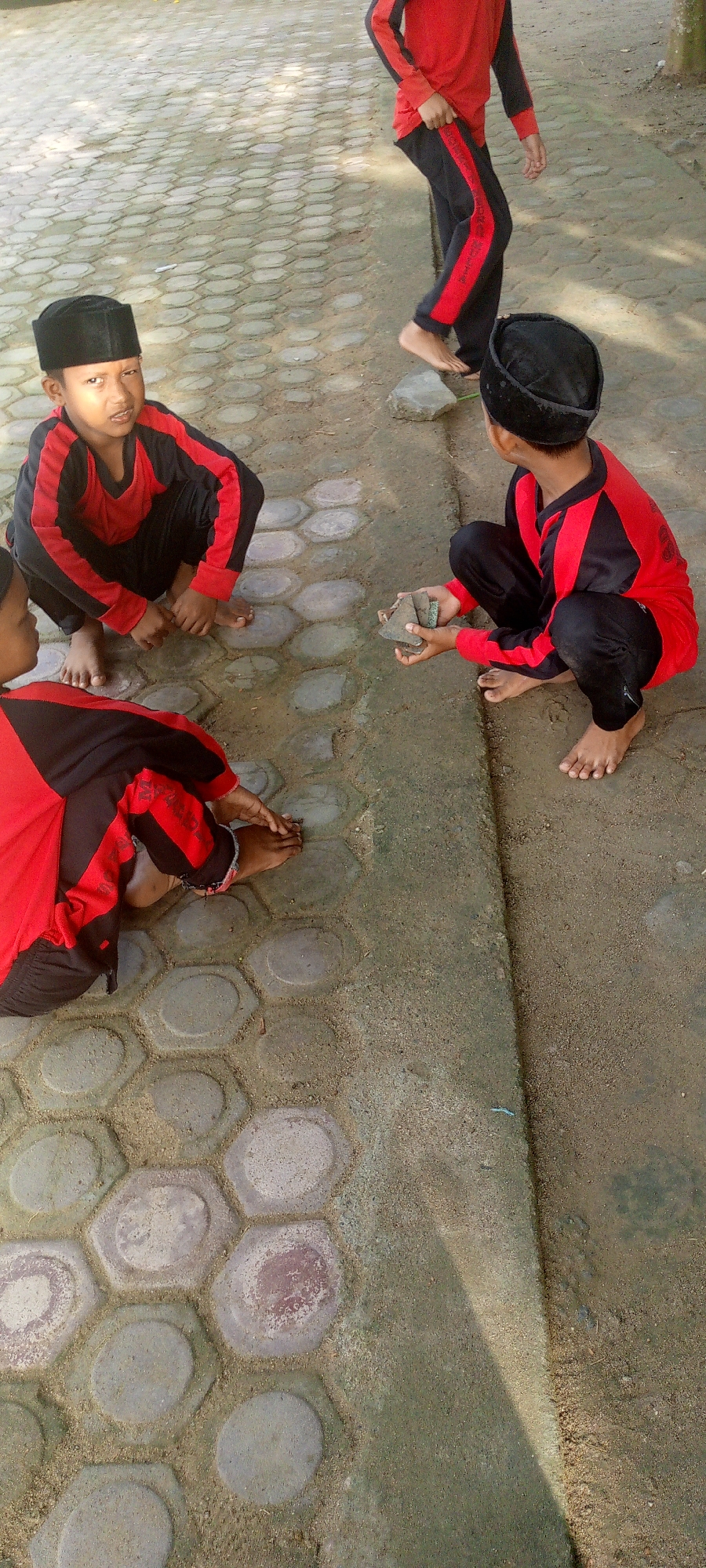 In Baratpayaitek, Indonesia, four young boys, dressed in matching red and black long-sleeved shirts and black pants, are engaged in an outdoor activity on a paved area with a dirt border. Two boys are in the foreground, one on the left looking towards the camera with a neutral expression, and another in the center, crouched with his back partially to the viewer, holding a small object. Two other boys are further in the background. One, on the right, is also crouched and holding a small object in his hands. The other boy, on the far right, is standing and walking away, with his bare feet visible. The ground is a mixture of hexagonal paving stones and packed dirt, with scattered small stones and sand. The lighting suggests it is daytime, with some shadows cast on the ground.