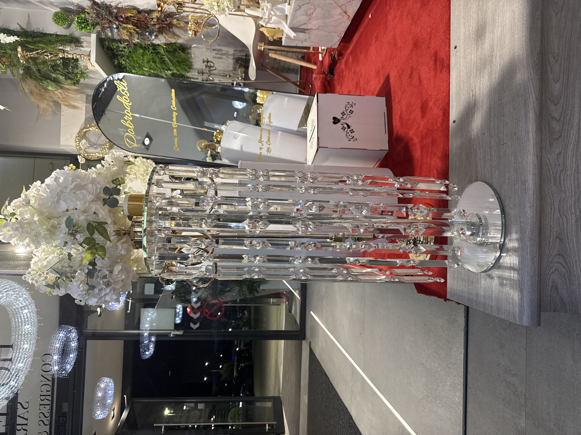The image shows a store window display in Ilidža, Bosnia and Herzegovina. The display features a tall, crystal-covered stand adorned with white flowers, and a black, arched mirror with text in gold. Behind the display, the interior of the store is visible, with shelves filled with decorative items, including plants, gold accents, and a white chair. A red carpet covers the floor in front of the display. The entrance to the store is also visible, with glass doors and reflections of chandeliers and lights. The scene appears to be set up for a special event, possibly a celebration or a wedding.