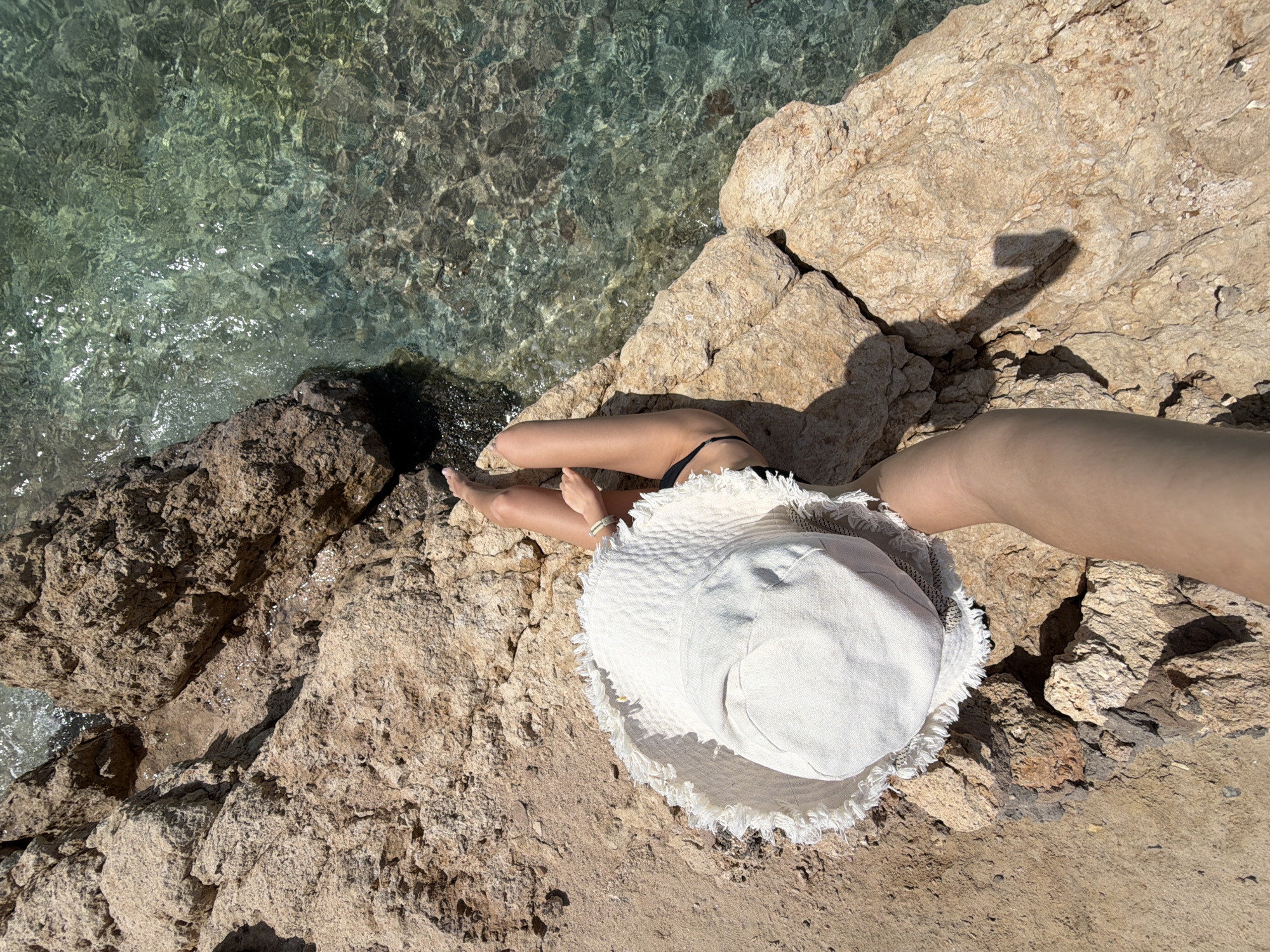 A top-down shot captures a person sitting on a rocky beach beside clear, turquoise water, likely in Sharm El Sheikh, Egypt. The person is wearing a white, fringed sun hat and a black bikini bottom. Their feet dangle over the edge of the rocks towards the water. The rocks are light tan and vary in size and texture. The water is clear enough to see the rocks beneath its surface. The shadow of the camera is cast on the right side of the rocks.