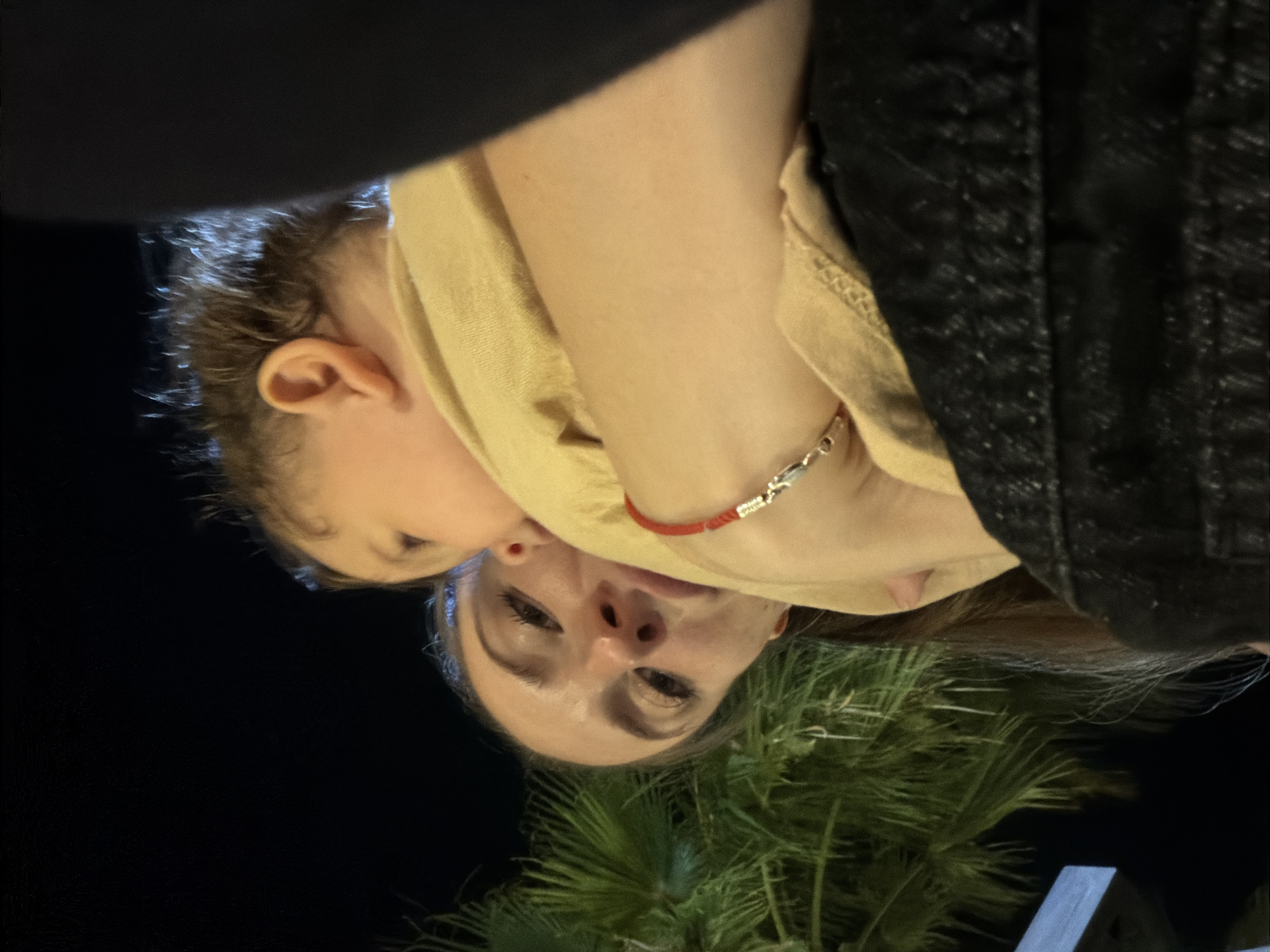 A young woman, with long brown hair and light-colored eyes, holds a baby in her arms. The woman is wearing a dark-colored jacket. The baby, with light brown hair, is looking away from the camera. A red bracelet is visible on the woman's wrist. The background features palm trees and a dark, nighttime sky, suggesting an outdoor location. The image appears to have been taken in Sharm El Sheikh, Egypt, a popular tourist destination known for its palm trees and nighttime ambiance. The lighting suggests it is evening or night.