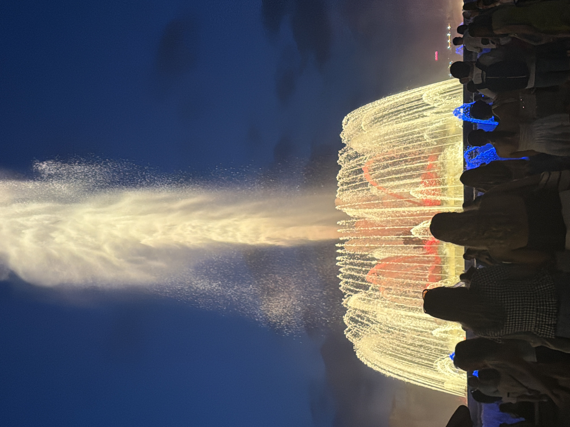 The image depicts a night scene at the Magic Fountain of Montjuïc in Barcelona, Spain. A large, illuminated fountain is in the foreground, with water jets reaching high into the dark blue sky. The fountain is lit with warm yellow and red lights, creating a striking visual effect. In front of the fountain, a crowd of people are gathered, silhouetted against the bright lights and water. The people are facing the fountain, seemingly watching the display. The atmosphere appears festive, with many individuals enjoying the spectacle. The sky is a deep, clear blue, indicating it is nighttime. The overall scene captures the beauty and grandeur of the fountain show.
