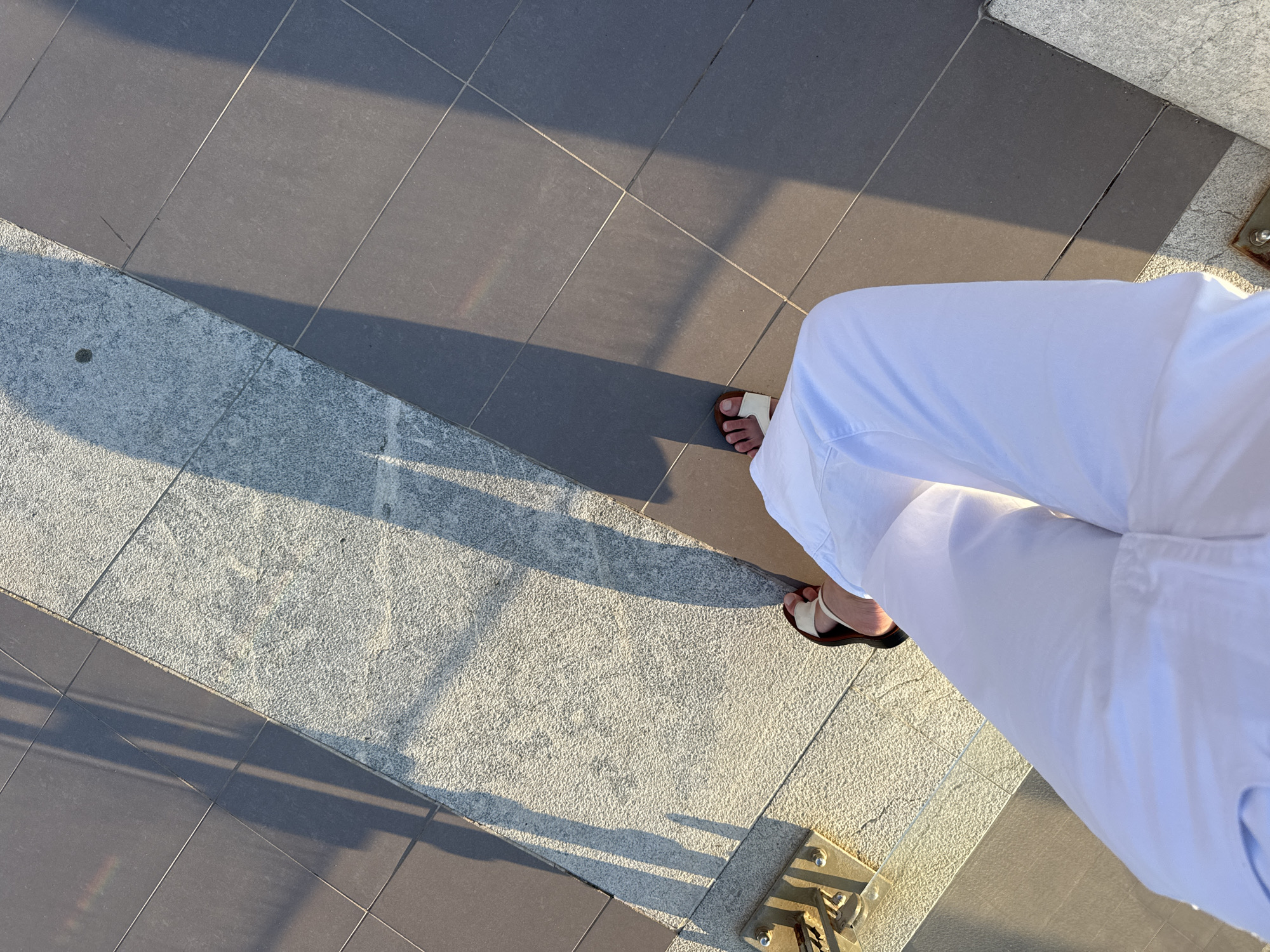 In Capaci, Italy, a first-person perspective shot captures a person wearing white pants and sandals, standing on a tiled walkway. The sun casts long shadows from an unseen structure, creating a striped pattern across the ground. The walkway is composed of two distinct surfaces: a textured, light-colored stone area and a darker, smooth tiled section. A small, faint rainbow-like spectrum is visible on the darker tiles. The image focuses on the ground, with the person's legs and feet occupying the lower portion.