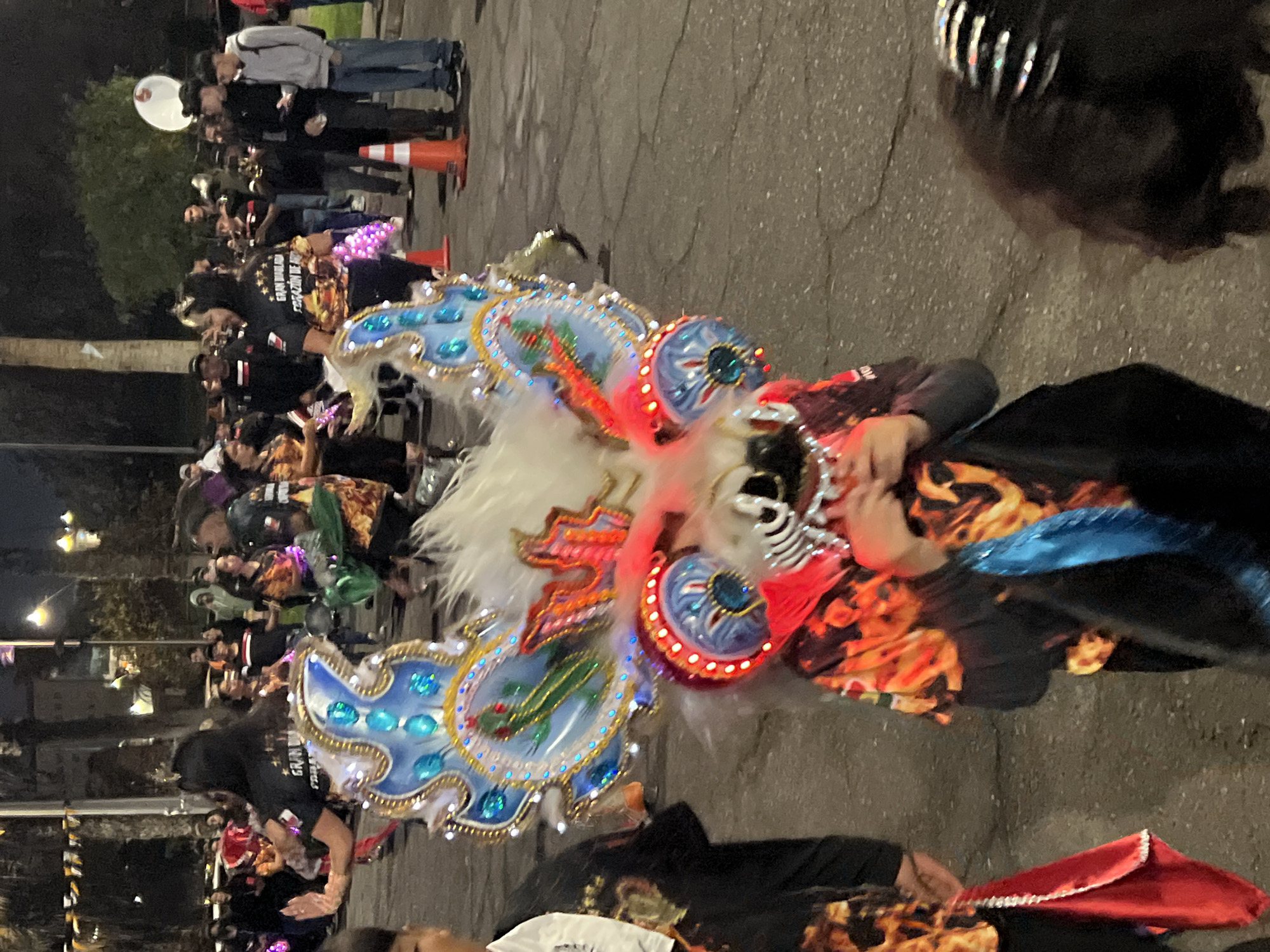 In Arica, Chile, a nighttime procession features individuals in vibrant costumes. The focal point is a person wearing an elaborate mask and headdress, adorned with blue, red, and white embellishments, and illuminated by red and blue lights. The mask has a skeletal appearance with glowing red accents around the eyes and mouth. The headdress, styled with white fur and large, brightly colored wing-like structures, features a depiction of a green cactus within a blue circular emblem. The individual's attire includes a black garment with a fiery orange and yellow print. To the left, another person wears a black shirt with white lettering and a red draped cloth. Behind them, several onlookers are visible, some holding illuminated objects. In the background, palm trees and streetlights indicate an urban setting. Orange traffic cones are placed along the paved street, suggesting a closed-off area for the event.