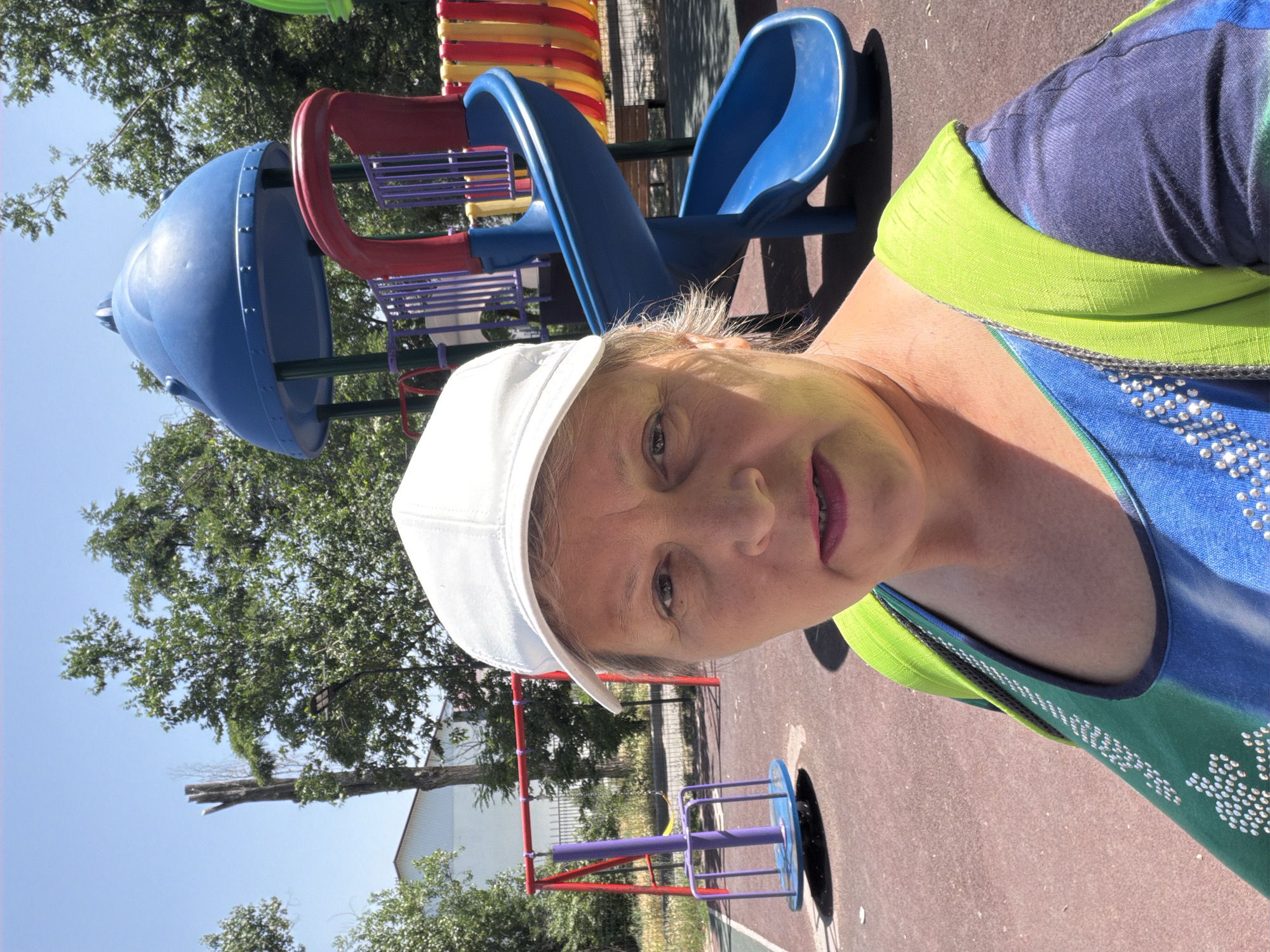 A middle-aged woman with fair skin and graying blonde hair, wearing a white baseball cap, a green and blue patterned top adorned with rhinestones, and a bright lime green scarf draped over her shoulders, poses for a selfie on a playground. The playground features a blue spiral slide, a red and yellow panel fence, a red swing set with a yellow seat and a purple carousel. The ground is a reddish-brown, soft surface typical of playgrounds. In the background, lush green trees and a building with a white facade are visible under a clear blue sky. A dead, splintered tree trunk stands prominently on the left side of the frame, with a black lamppost nearby. The lighting suggests it is daytime, likely a sunny afternoon.