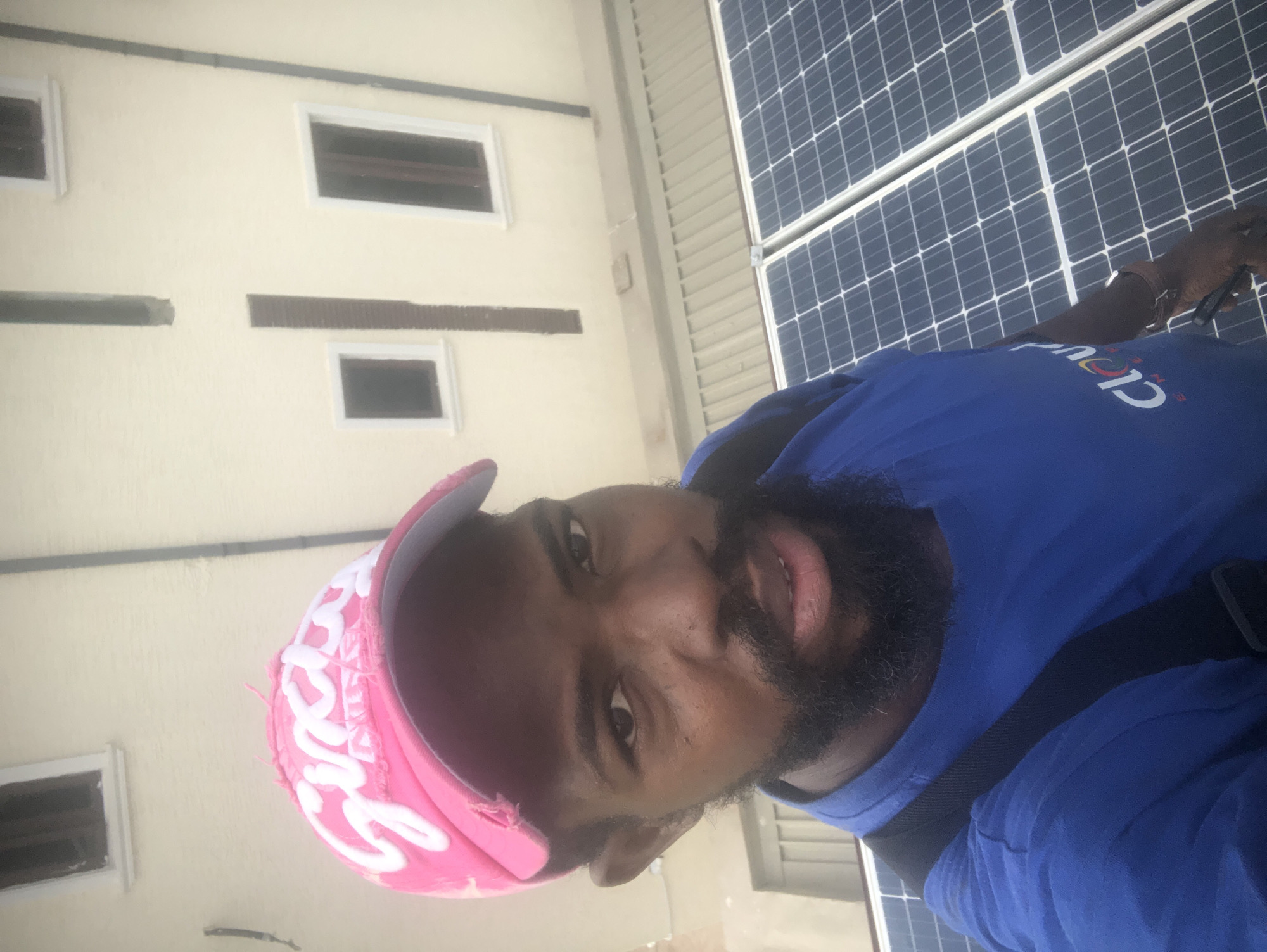 A medium shot captures a Black man in a bright pink cap and blue shirt standing on a roof with solar panels in Ilado, Nigeria. He is looking directly at the camera, and his face is partially obscured by the brim of his cap. The background shows a light-colored building with windows. The man appears to be working, as evidenced by the solar panels, and his attire suggests he might be an electrician or solar panel installer. The image is taken from a high angle, looking down at the man and the surrounding area. The lighting is bright, indicating it is daytime.