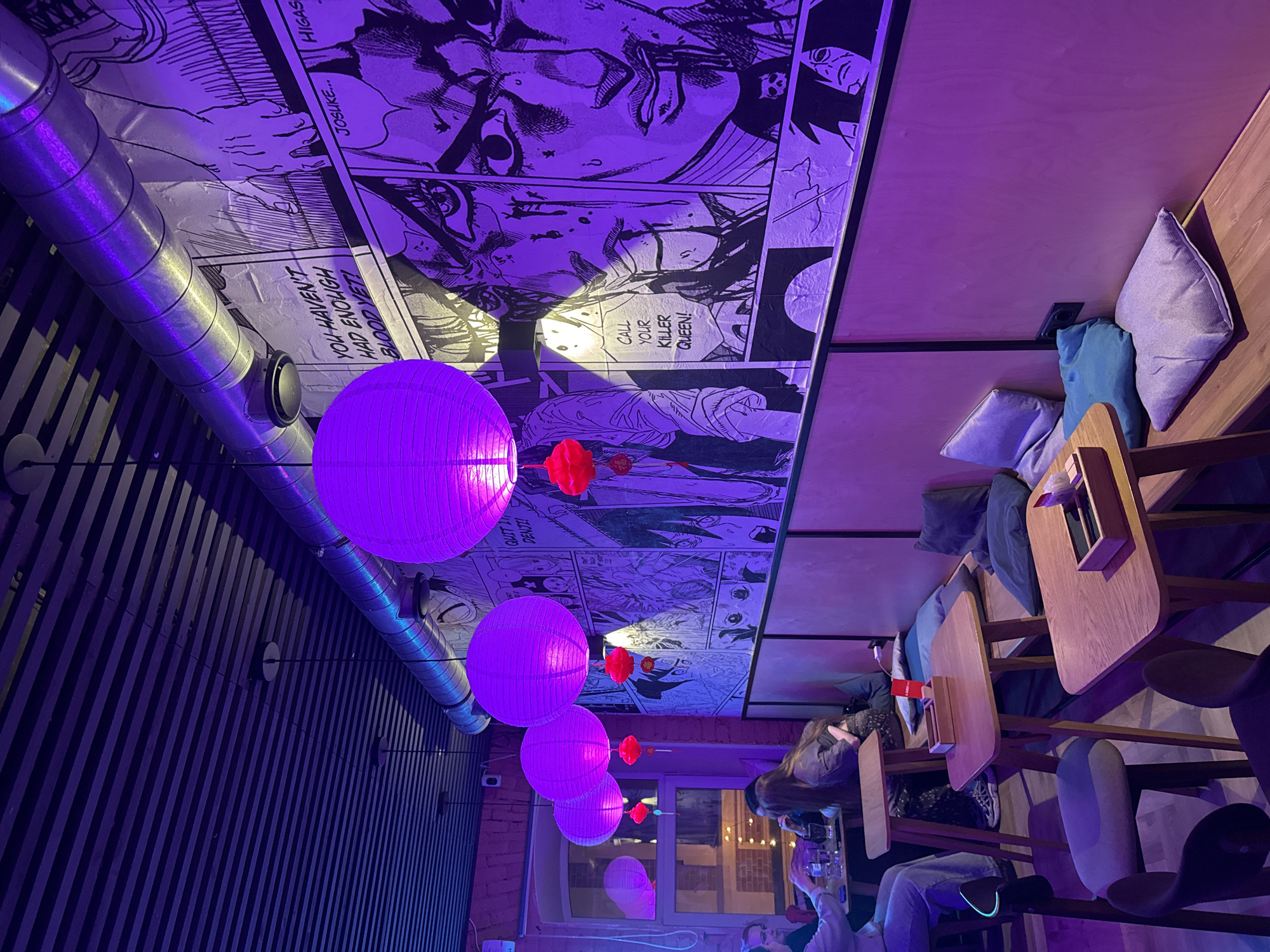 The image shows the interior of a room with a distinctive aesthetic. The ceiling is decorated with black and white manga panels and has several purple spherical lanterns hanging from it. The room features dark, vertical striped walls and a metal ventilation pipe. On the right side of the image, there are wooden tables and chairs with cushions, where several people are seated and engaged in activities such as dining and drinking. The lighting in the room is predominantly purple, which casts a cool tone over the scene. The room appears to be a restaurant or bar, based on the seating arrangement and the presence of drinks and tables.