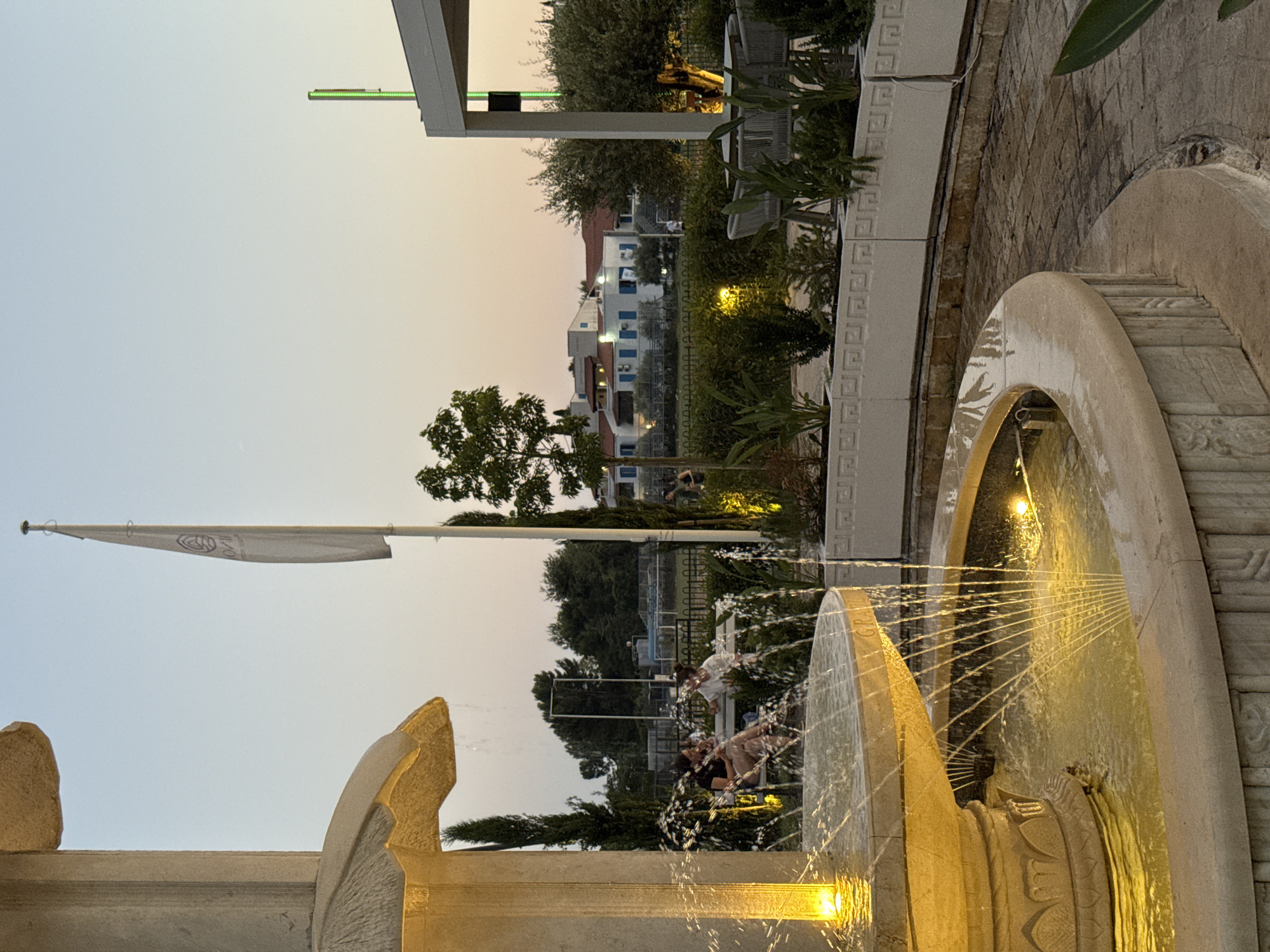 The image depicts an outdoor scene at dusk, likely in a resort or public area with a fountain in the foreground. The fountain, made of light-colored stone, has multiple tiers and is illuminated by yellow lights, with water spraying outwards. Two women are seated on a bench in the middle ground, appearing to be in conversation. Beyond them, a white building with blue shutters and red-tiled roofs is visible, surrounded by greenery and trees. A flag with a logo is planted on a flagpole to the left of the building, and a modern structure with a white canopy and a green light fixture is to the right. The sky is a soft gradient of orange and blue, indicating sunset. This setting is consistent with the waterfront areas or hotel complexes found in Budva, Montenegro, a popular tourist destination known for its beaches and historical sites.