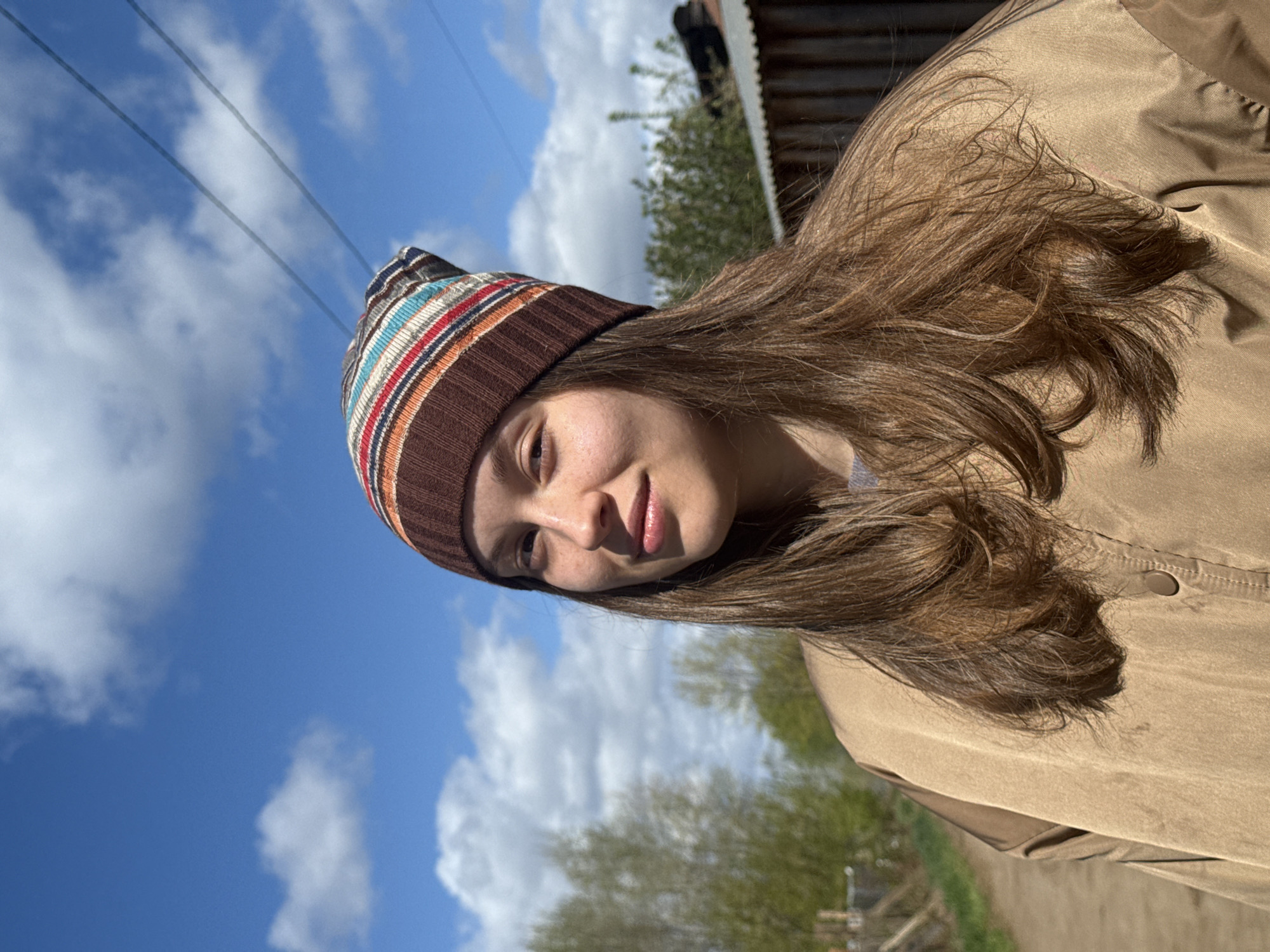 The image depicts a woman wearing a brown and multi-colored striped beanie and a tan jacket. She is facing slightly to the left with a gentle smile. The background includes a clear blue sky with scattered white clouds, and elements that suggest an outdoor setting. There's also a glimpse of power lines in the sky and hints of greenery, possibly trees, and a building roof in the background. The photo appears to have been taken on a sunny day. Location specifics beyond "outdoors" or "Russia" cannot be determined.