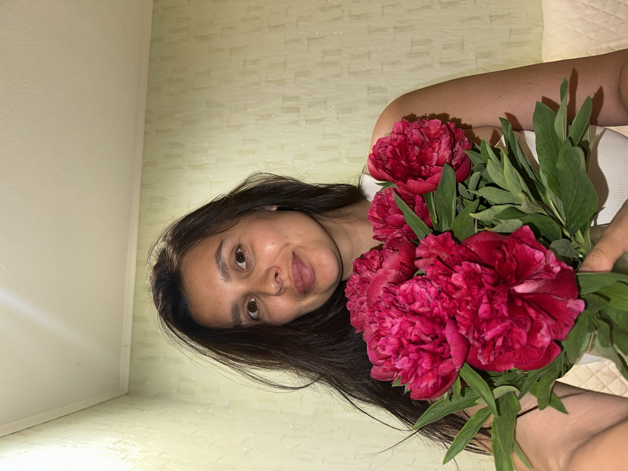 A woman with long, dark hair is holding a bouquet of dark pink peonies. She is wearing a white top. The background consists of a textured beige wall. It is not possible to verify the location as Chaikovsky, Russia, from the image.