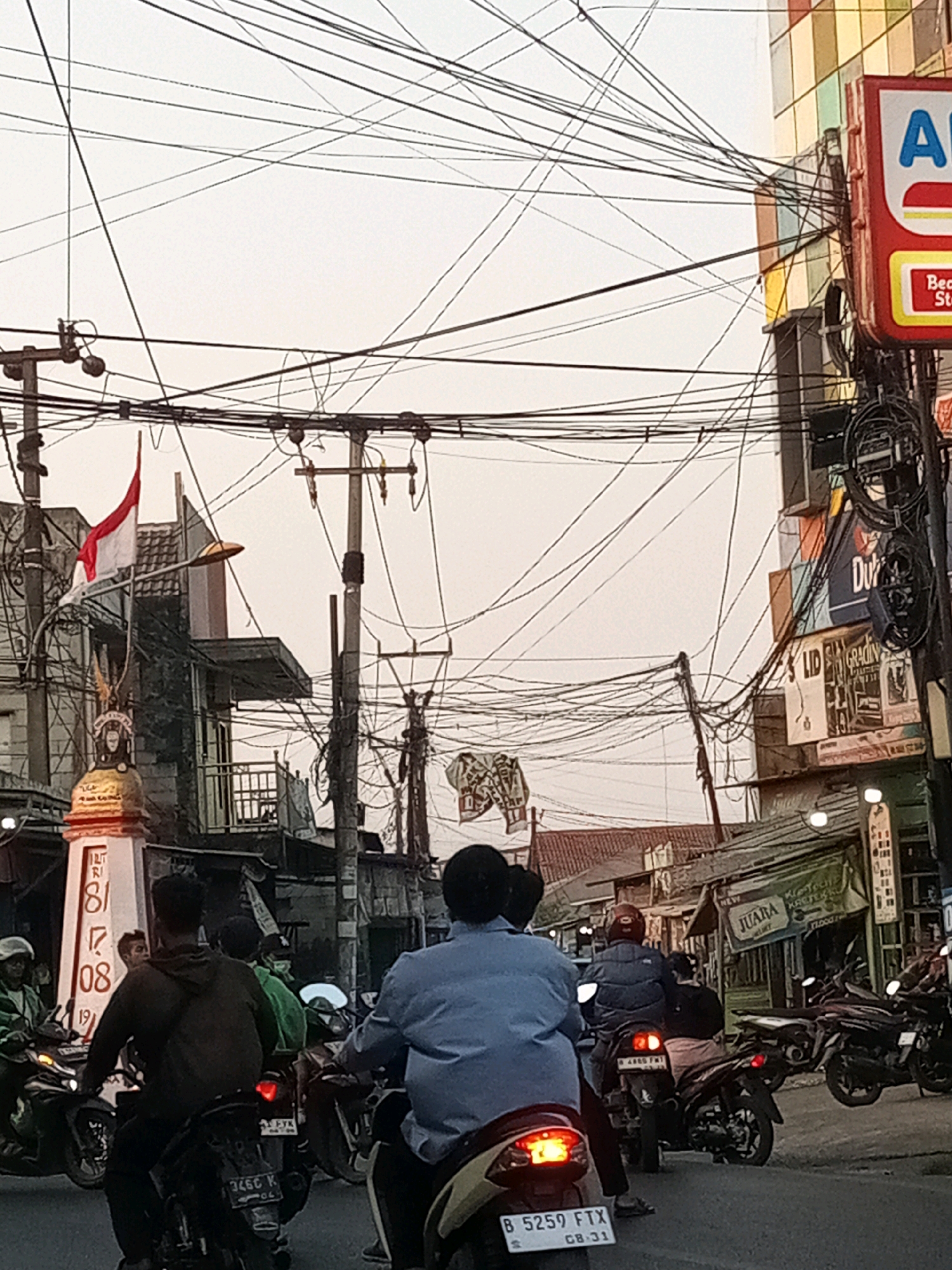 The image depicts a street scene in Karangsatria, Indonesia, characterized by dense overhead wiring. In the foreground, several motorcycles and their riders are visible. One motorcycle has a license plate reading "B 5259 FTX 08-31". To the left, a monument or pillar with the numbers "181 08 19" is present, topped with a golden statue. A red and white Indonesian flag is attached to a pole near the monument. Buildings line the street, some appearing to be shops with signage. One prominent sign on the right displays "Al". The sky is overcast.