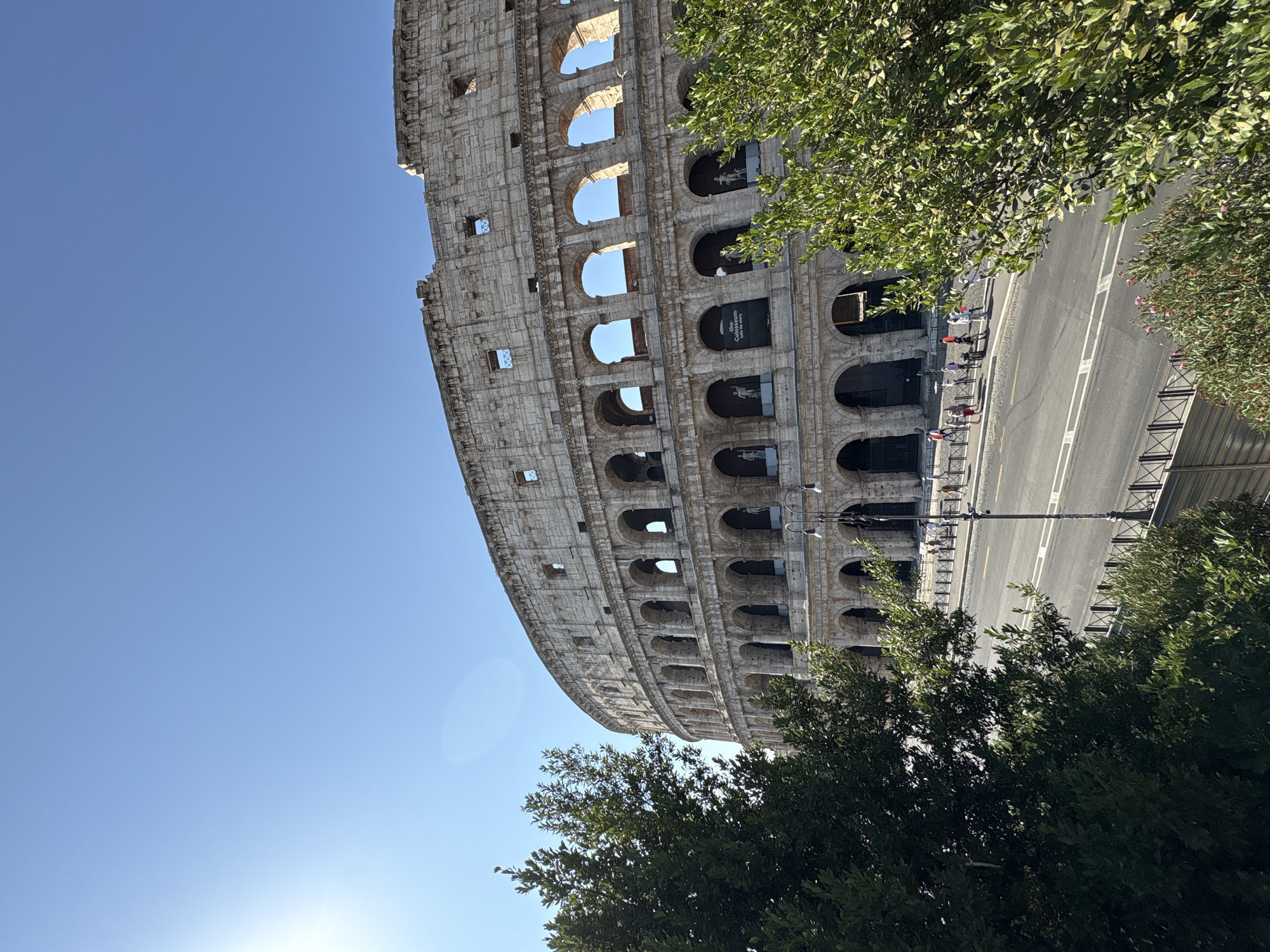 This is a full outdoor, eye-level shot of the Colosseum in Rome, Italy, on a bright, clear day. The sun is shining, casting a bright light on the ancient amphitheater. The Colosseum, a massive oval structure made of stone, dominates the upper right portion of the frame. Its arched windows and weathered facade are clearly visible. Lush green trees frame the left and right sides of the image, with their branches partially obscuring the view of the Colosseum. In front of the Colosseum, a paved road runs horizontally across the lower half of the image, with a sidewalk along its edge. A few people are seen walking on the sidewalk, some appear to be tourists. A street light stands on the sidewalk. The sky is a clear, bright blue with no clouds. The overall scene is one of historical grandeur set against a vibrant natural backdrop.