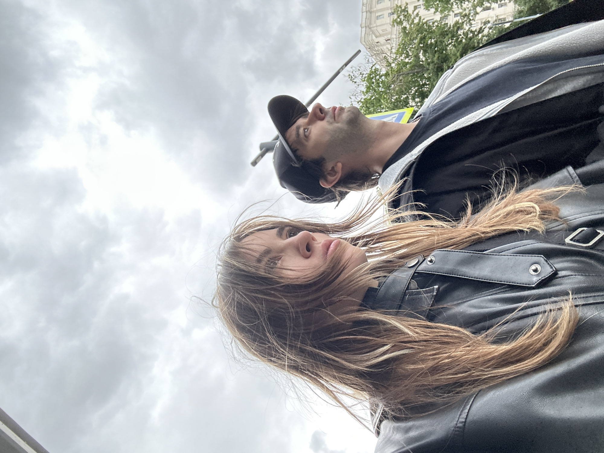 A medium shot captures a young woman and a man from the chest up, looking to their right under a cloudy sky. The woman, on the left, has long, wavy brown hair that is windswept. She is wearing a black leather jacket. The man, on the right, is wearing a black baseball cap, a black t-shirt, and a grey zip-up hoodie. Behind them, a light-colored building with visible windows is partially obscured by green trees. The scene appears to be outdoors in an urban setting, possibly a street in Moscow, Russia, given the architectural style of the building. The lighting is diffused due to the overcast conditions.