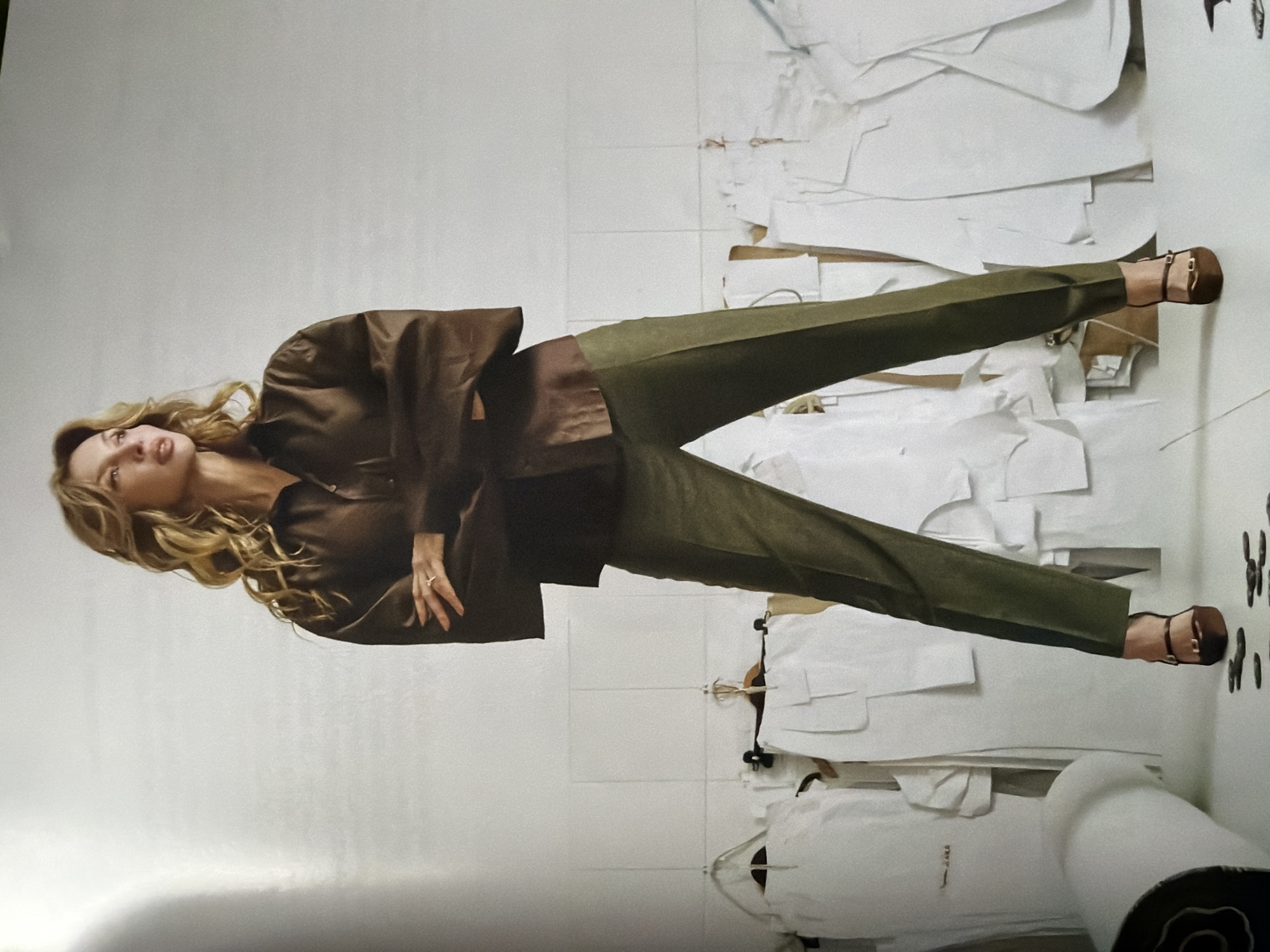A full-length studio shot captures a slender woman with long blonde hair, lying sideways in a brown blouse, green pants, and brown sandals. She is in a room against a white tile wall with rolls of white paper containing cutouts of various shapes. The city of Moscow, Russia is not discernible in this image.