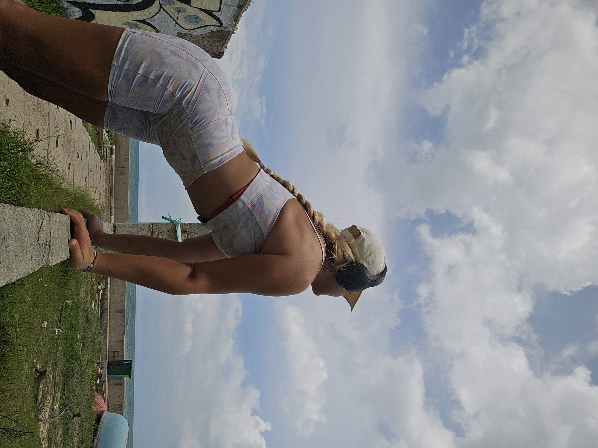 A woman is performing a yoga pose outdoors with the ocean and sky in the background. She is wearing a patterned sports bra and matching shorts, a beige baseball cap, and black headphones. Her blonde hair is styled in a braid that hangs down her back. Her arms are extended, hands flat on a concrete ledge, and her body is in a low plank position. The scene is set against a cloudy blue sky, and the ocean is visible in the distance. The sun appears to be shining, suggesting it is daytime. The woman's posture and focused expression suggest concentration and dedication to her exercise. There is graffiti on a wall behind her, and some green grass and debris are visible on the ground in the foreground. A green trash bin and a blue object are also present in the scene near the water.