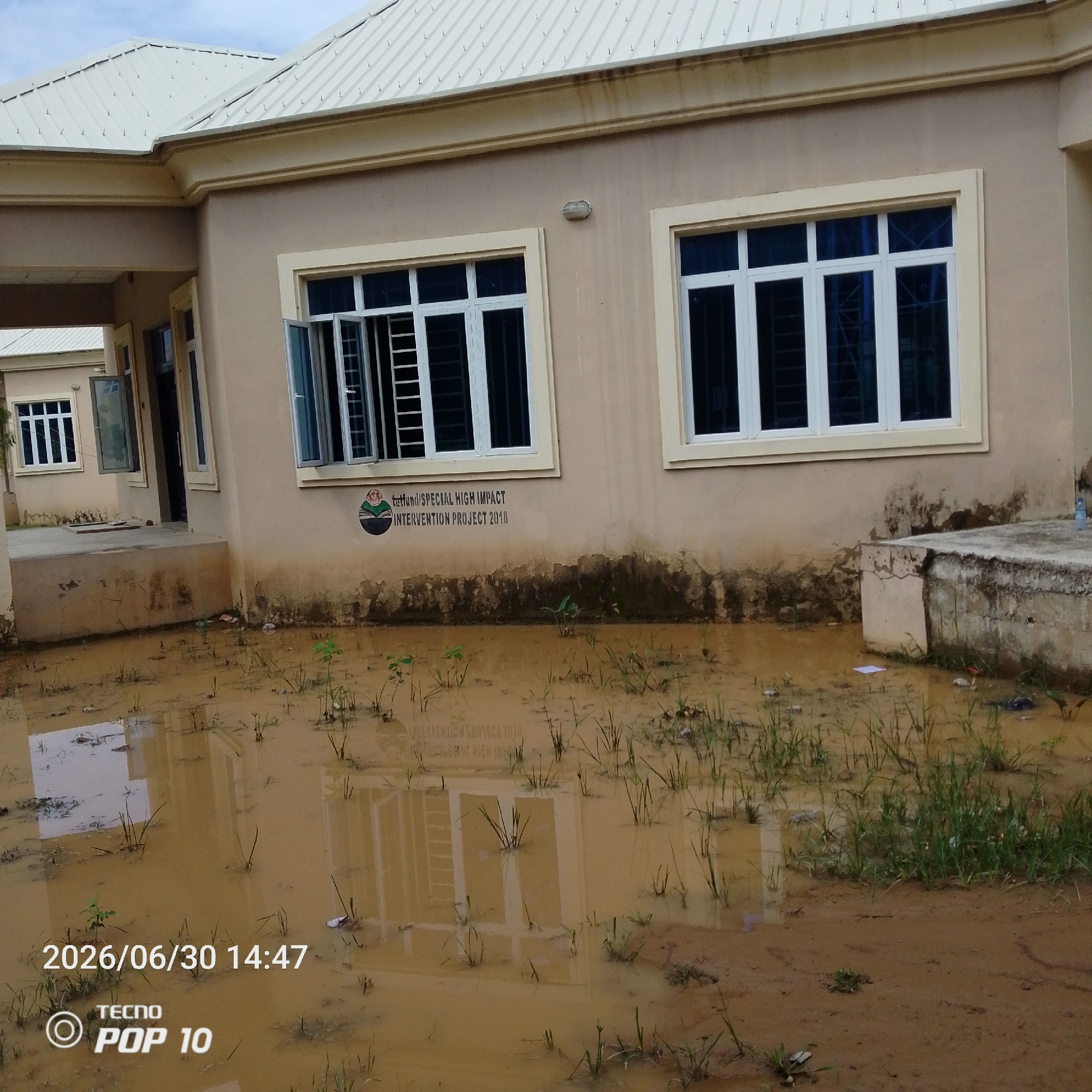 The image shows a flooded area in front of a light-colored, single-story building with a white metal roof. The building has several windows with white frames and blue-tinted glass. The front of the building is partially obscured by murky floodwater. Small green plants are growing out of the water and mud.
On the wall of the building, there is a circular logo with an image of a person reading a book, and text that appears to read "tethfundSPECIAL HIGH IMPACT INTERVENTION PROJECT 2018".
The foreground is dominated by the brown, muddy floodwater with reflections of the building and sky. Some debris, including small pieces of plastic and paper, are visible in the water.
In the bottom left corner of the image, there is a timestamp "2026/06/30 14:47" and a "TECNO POP 10" watermark.
The scene suggests a recent rainfall event or a natural disaster like flooding. The sky is overcast, indicating potentially rainy or post-rainy weather. The setting appears to be a semi-urban or rural area in Jalingo, Nigeria, given the context. No people are visible in the image. The overall impression is one of an unfortunate situation where a building and its surroundings have been inundated with water.
