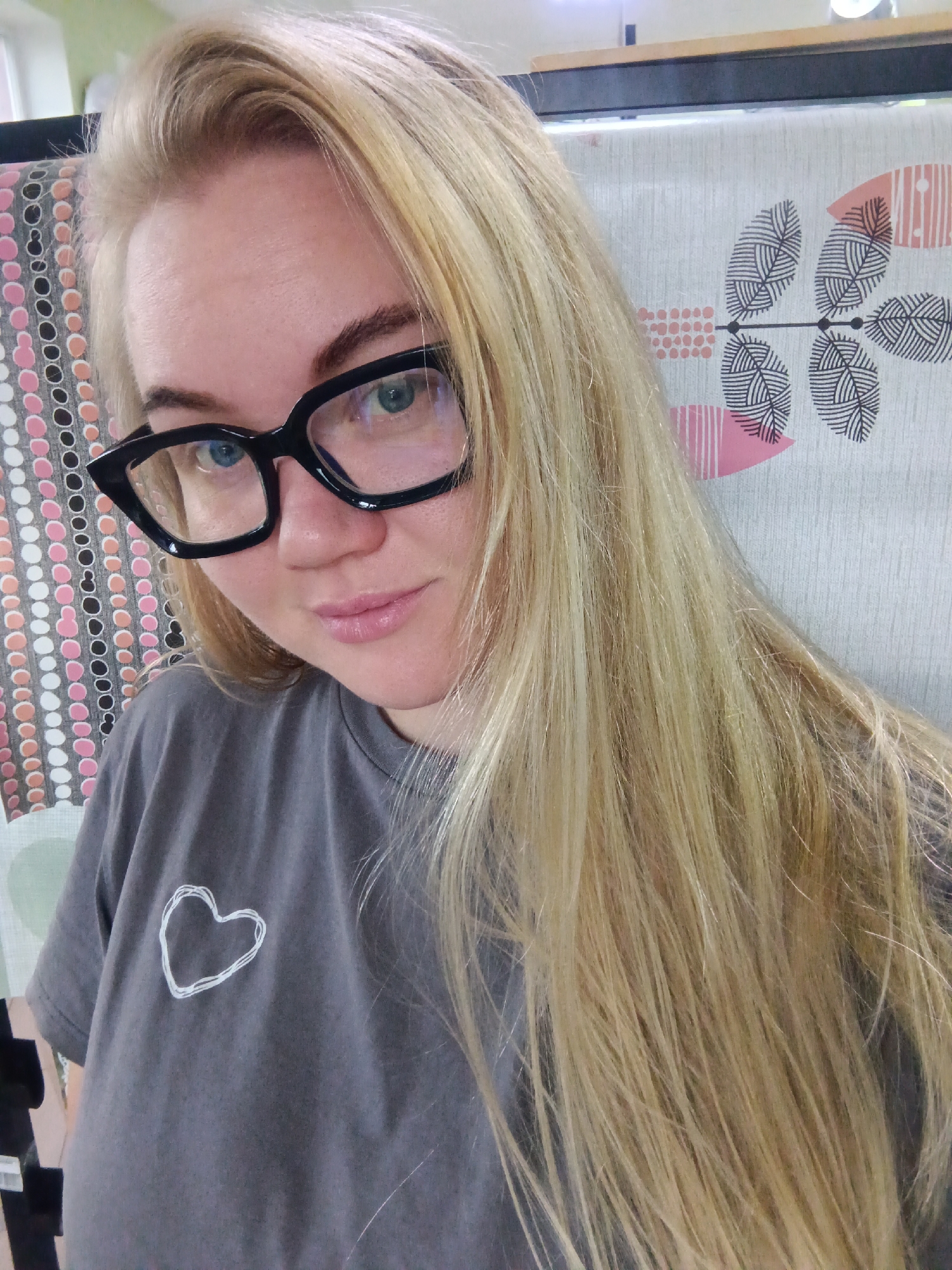 A woman with blonde hair and blue eyes is pictured wearing a gray t-shirt with a white heart design and black cat-eye glasses. She is indoors, with a patterned background behind her. The image's lighting is natural. The scene appears to be a casual selfie taken within a residential setting in the city of Fokino, Russia.