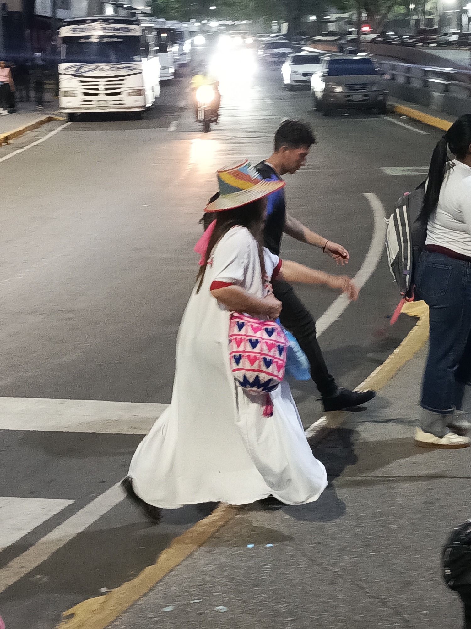 The image captures a nighttime scene on an urban street, likely an intersection or crosswalk. In the foreground, three individuals are walking across the asphalt road from left to right.
The central figure is a woman wearing a long, flowing white dress with red trim on the sleeves. She has long dark hair and a colorful, woven hat with stripes of blue, red, and yellow. She carries a distinctively patterned bag, possibly woven, featuring pink, blue, and white geometric designs resembling hearts or triangles. To her right, a man with dark hair walks beside her, dressed in a dark t-shirt and dark pants. His right hand is outstretched, as if reaching or gesturing. Further to the right, partially visible, is another person wearing a white top, blue jeans, and carrying a black and white striped backpack.
The road features white painted crosswalk lines and a yellow dividing line. In the background, several vehicles are present. A large white bus with "Piso" written on its front grille is prominent on the left side of the road, appearing to be in motion with some motion blur. To the right of the bus, a bright headlight indicates a motorcycle also moving. Further down the road, on the right side, other vehicles, including a dark car, are visible with their headlights on, indicating it is dark outside. The artificial lighting from streetlights and vehicle headlights creates bright reflections and glares on the road surface. The overall impression is of a busy urban environment after dusk, with people navigating pedestrian crossings amidst traffic.