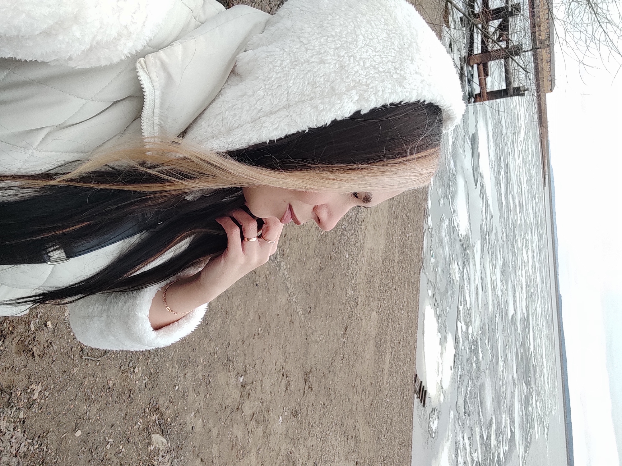 An outdoor portrait shot in Chaikovsky, Russia, features a person, likely female, depicted in profile, gazing downwards and slightly to her right. She has long, dark hair with prominent blonde highlights around her face and is seen gently holding strands of her hair with her right hand. She is wearing a thick, white, quilted winter jacket, distinguished by a fluffy, white hood and matching furry cuffs. On her right hand, multiple rings are visible, and her left wrist sports a delicate gold chain bracelet. Her lips are slightly parted, and her expression appears calm and thoughtful.
The setting suggests a cold climate, likely winter or early spring. The ground in the foreground is a rough, unpaved surface of dirt and small stones. To the person's right, an expanse of water is visible, covered in large, broken ice floes, indicating a frozen or recently thawed body of water, possibly a river or reservoir. In the background, beyond the icy water, dark, skeletal structures, which might be industrial remnants or an old pier, stand against a bright, overcast sky, accompanied by bare tree branches. The time of day is daylight. No visible text is present in the image.