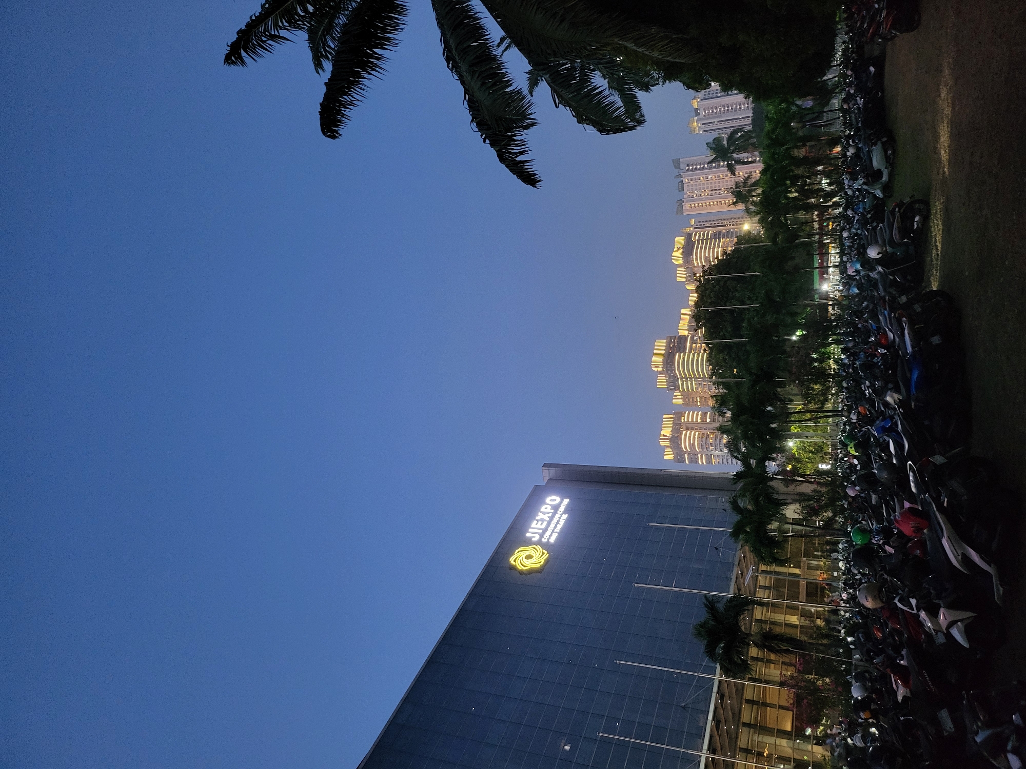 This media file captures an outdoor scene at dusk in North Jakarta, Indonesia. The sky is a deep twilight blue, with the last light of the day illuminating the windows of several tall buildings in the background. These buildings appear to be illuminated with warm, golden lights. In the foreground, a modern glass-fronted building with the illuminated text "JIEXPO CONVENTION CENTRE AND THEATRE" and a distinctive yellow logo is visible. Adjacent to the building, a large number of motorcycles are parked in rows on a grassy area, suggesting a popular event or gathering. Palm trees and other foliage are present, adding to the tropical setting. The overall atmosphere is calm and peaceful, transitioning from day to night.
