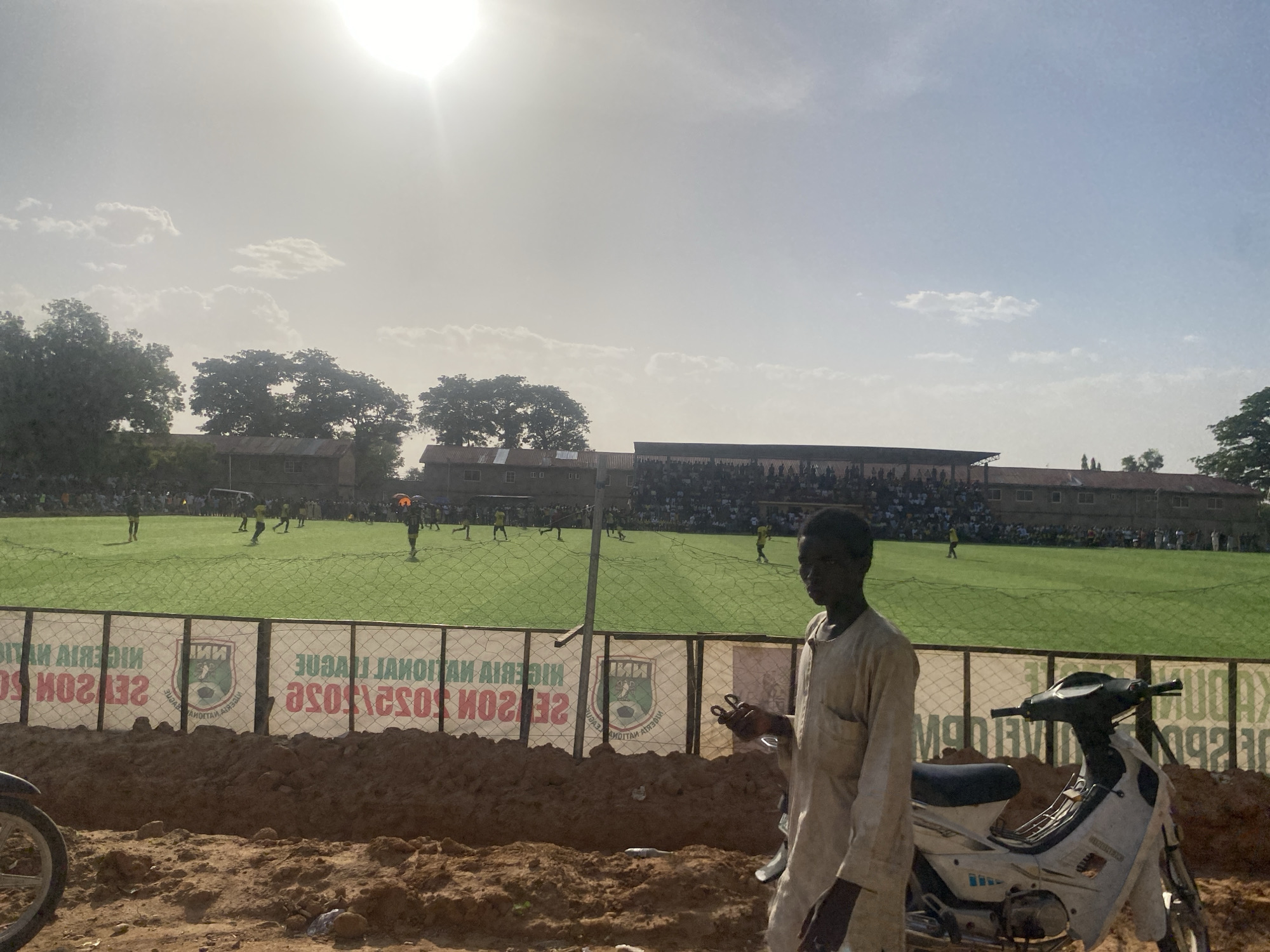 The image shows an outdoor scene in what appears to be Tudun Wada, Nigeria, with a soccer field in the background. A soccer game is in progress, with players in yellow and other colors visible on the green field. A crowd of spectators is seated in bleachers on the far side of the field. A fence with advertising banners, reading "NIGERIA NATIONAL LEAGUE SEASON" and others, separates the foreground from the soccer field.
In the foreground, a dirt area exists where the ground is uneven and sandy. A young man dressed in light-colored clothing walks by, holding an indiscernible object in his hand. Beside him is a parked motorcycle with a white and black color scheme. The sun is high in the sky, creating a bright, hazy environment. Trees and buildings are visible in the background beyond the soccer field.