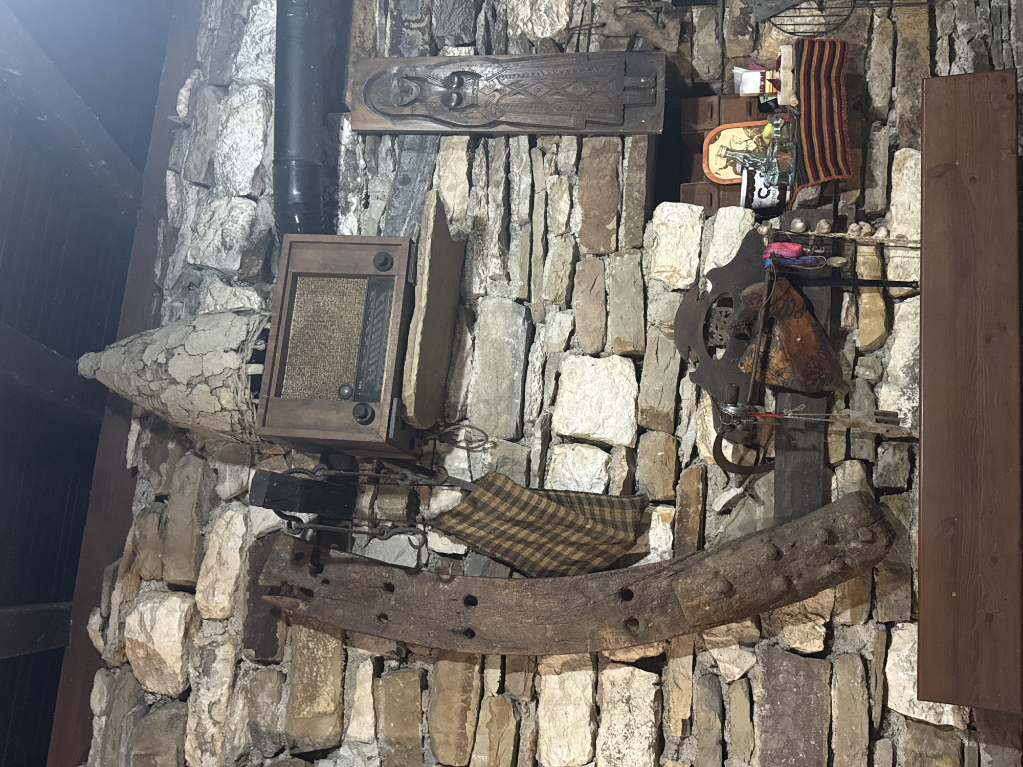 The image depicts a rustic interior wall constructed of rough-hewn stones. Mounted on the wall are various artifacts. To the left, a large, curved wooden object with holes is suspended by metal chains. Next to it hangs a checkered fabric. Above these items, a vintage radio sits on a wooden shelf. To the right of the radio, a carved wooden relief of a standing female figure is mounted on the stone wall. Below the radio and relief, a wooden beam extends horizontally, supporting a saddle-like object with decorative metalwork and various small items, including strings of garlic. A small shelf to the right holds a colorful striped textile, a jar, a glass bottle, and other decorative objects. A wooden plank runs horizontally across the bottom of the frame, suggesting a mantelpiece or shelf. The overall impression is one of historical or folk art display. This scene is located within a building in the village of Petrevene, Bulgaria.