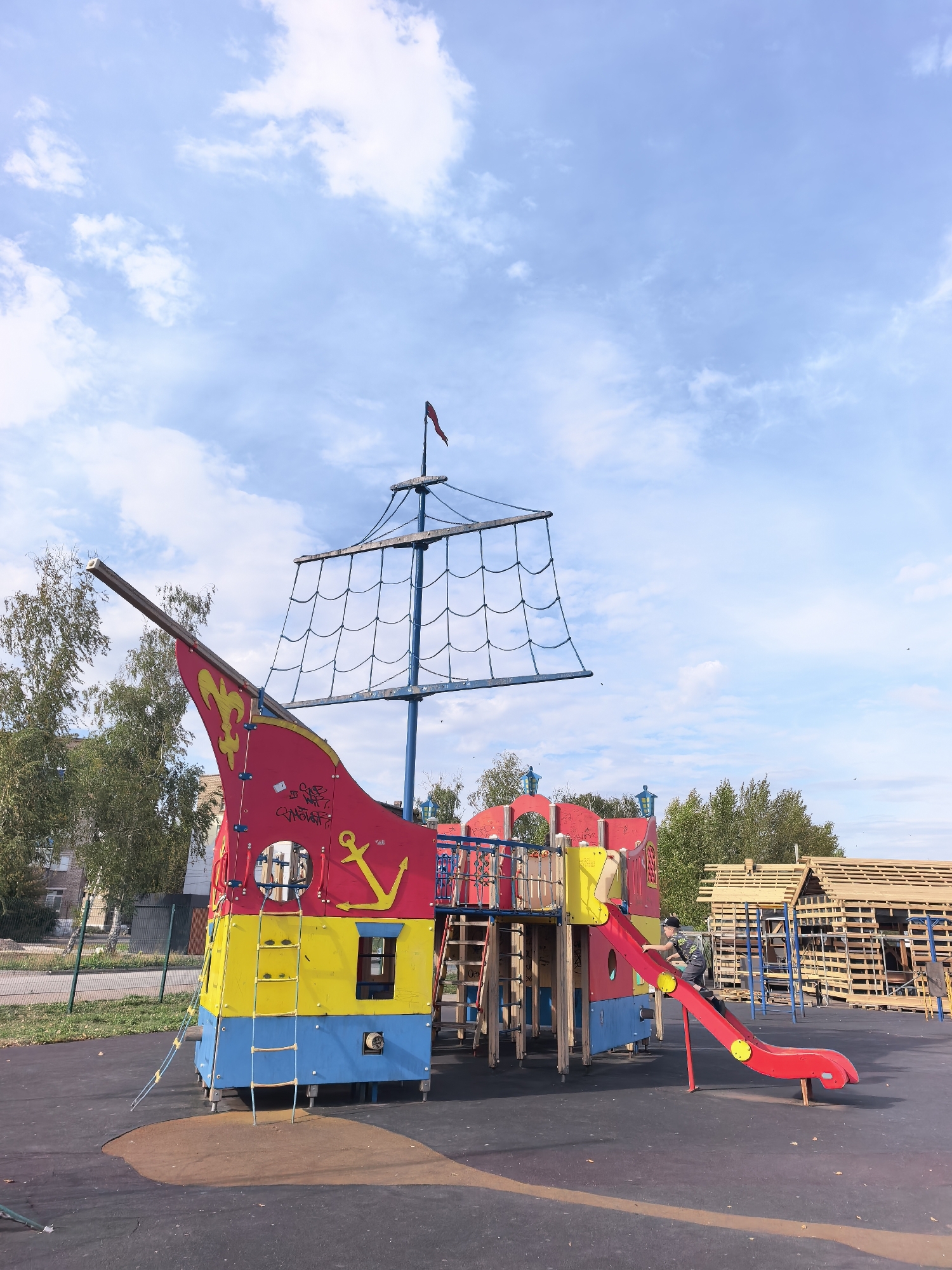 An outdoor playground in Sterlitamak, Russia, features a large, colorful pirate ship-themed play structure. The structure is primarily red, yellow, and blue, with a mast, rigging, and a red flag at the top. There's a red slide attached to the side, and a person is shown sliding down it. Steps and ladders are integrated into the play structure for access. In the background, there are trees and a partially constructed wooden structure that appears to be made of pallets. The ground is a dark, soft surface, likely rubberized safety flooring, with some scattered leaves. The sky above is blue with scattered white clouds.