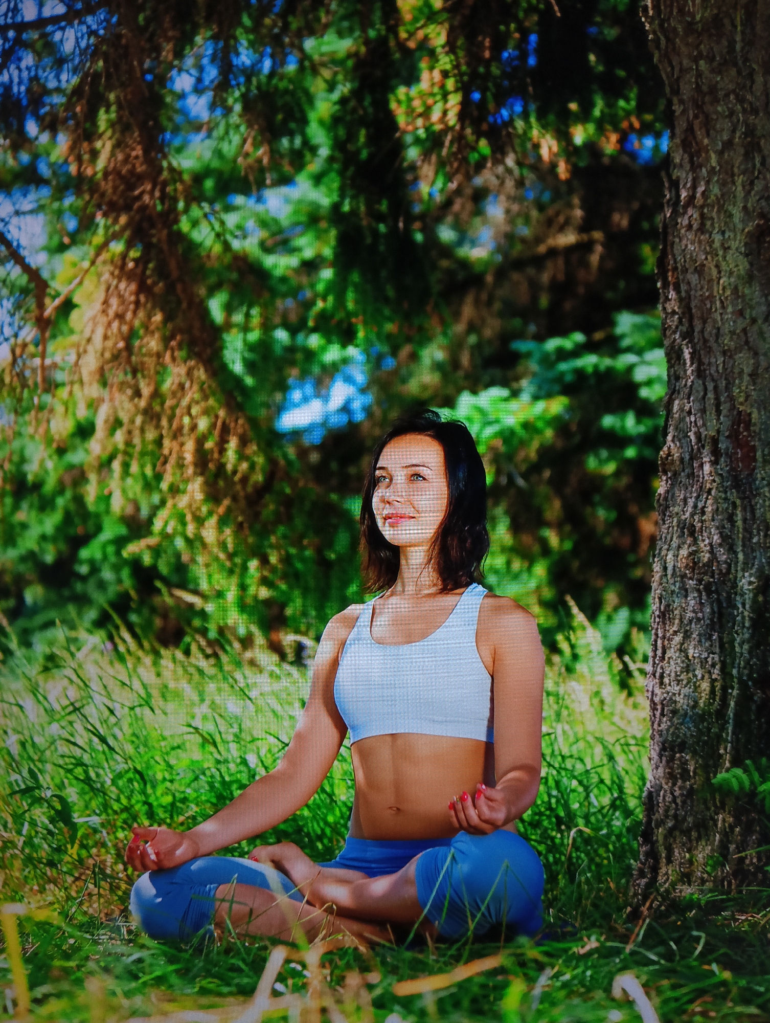 A woman is sitting in a meditative pose, likely lotus position, in a grassy area next to a tree. She is wearing a light-colored sports bra and blue leggings. Her dark hair is shoulder-length. The background is composed of trees and foliage, suggesting a natural outdoor setting. The specific location in Nha Trang Ward, Vietnam, cannot be definitively determined from the visual data; no landmarks or details provide that information.