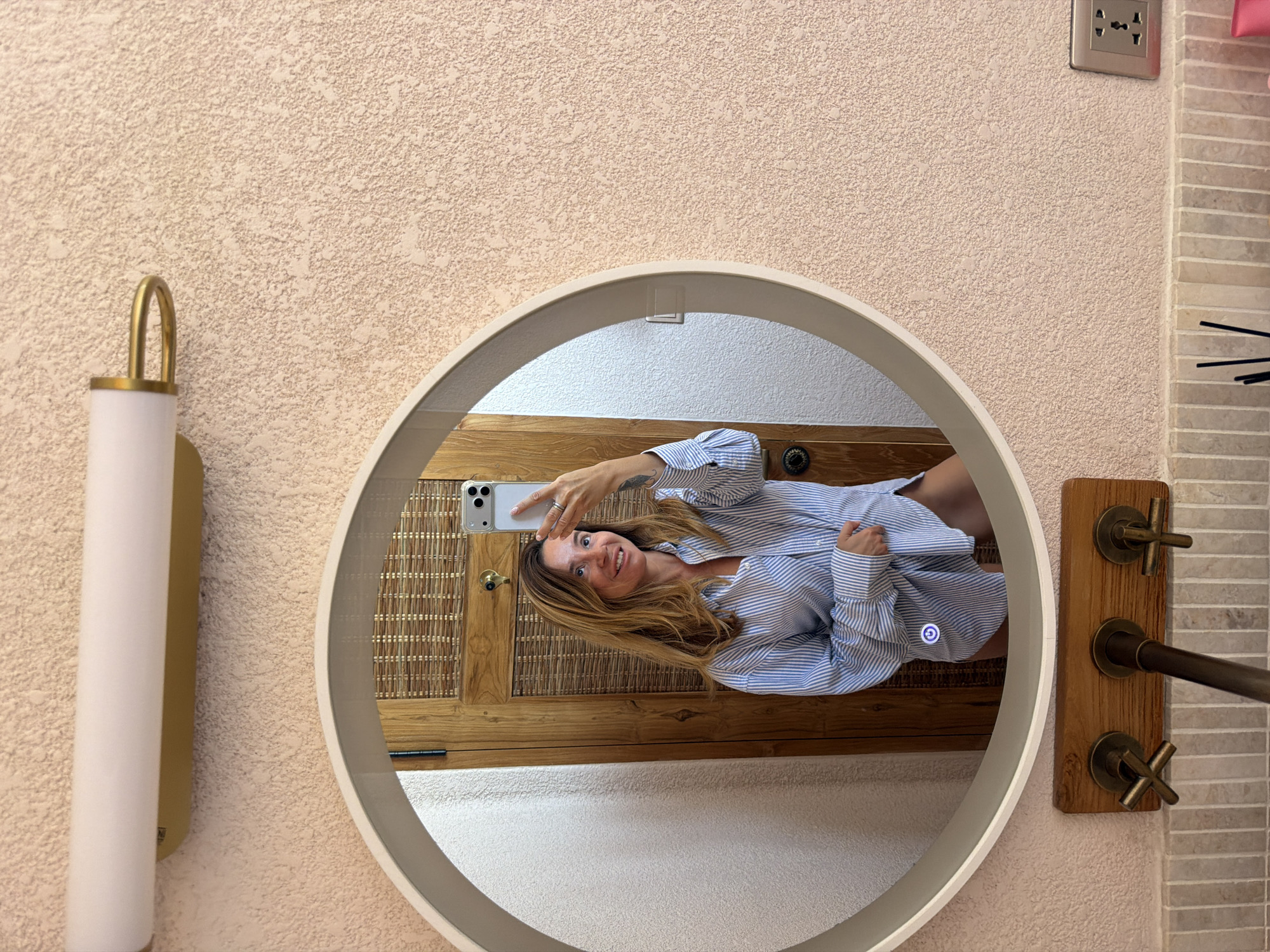 A woman with shoulder-length blonde hair, wearing a blue and white striped button-down shirt, takes a mirror selfie in a bathroom. She is holding a silver smartphone in her right hand. The mirror is circular with a white frame and is mounted on a textured, light pink wall. Behind her, a wooden door with woven panels is visible. To the left of the mirror is a white wall. Below the mirror, there is a wooden fixture with three brass faucets, set against a tiled backsplash. The overall scene appears to be in a residential bathroom, possibly in a hotel or resort, in Mengwi, Indonesia. The lighting suggests an indoor setting, and the overall aesthetic is modern and clean.