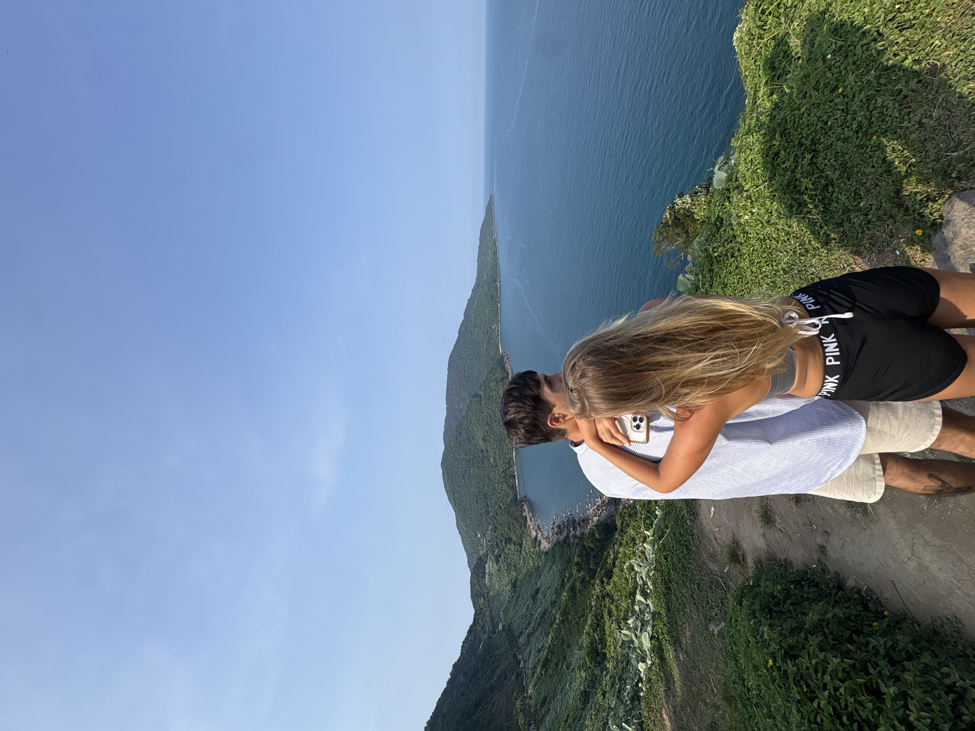 The image, taken on a clear, sunny day, captures a couple standing on a cliff overlooking a vast ocean and lush green mountains, characteristic of coastal areas like Đà Nẵng, Vietnam. The sky is a vibrant blue with minimal clouds, suggesting excellent weather conditions, likely late morning or afternoon given the bright sunlight.
The female subject, with long blonde hair flowing down her back, is seen from behind and slightly to the side. She wears a grey sports bra and black athletic shorts, on the waistband of which the word "PINK" is clearly visible in white. She holds a light-colored smartphone in her left hand and has her right arm gently around the male subject. The male, with dark hair, is partially visible from the side, wearing a light blue and white striped short-sleeved shirt and light beige shorts. The couple appears to be intimately enjoying the panoramic view.
The scene features a wide expanse of deep blue ocean meeting a long line of verdant green mountains and hills that stretch along the coastline. The cliffs where the couple stands are covered in dense green foliage, with hints of a dirt path nearby. The overall impression is one of a serene and picturesque moment shared by the couple amidst a stunning natural landscape.
