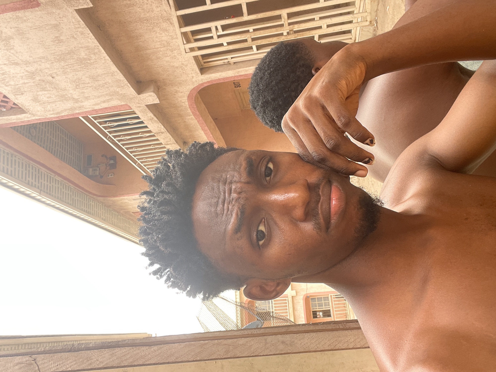 The image shows two Black men, presumably in Lokoja, Nigeria, based on the building style and complexion of the individuals. The photo is taken at an unusual angle, possibly from below. The closer man has short, dark, curly hair and a slight beard. He is shirtless. The other man is partially visible behind him and appears to have a similar hairstyle. Both are brown-skinned. The background consists of a multi-story building with window bars and some architectural detailing. The lighting suggests it is daytime. There is a wooden structure in the foreground.